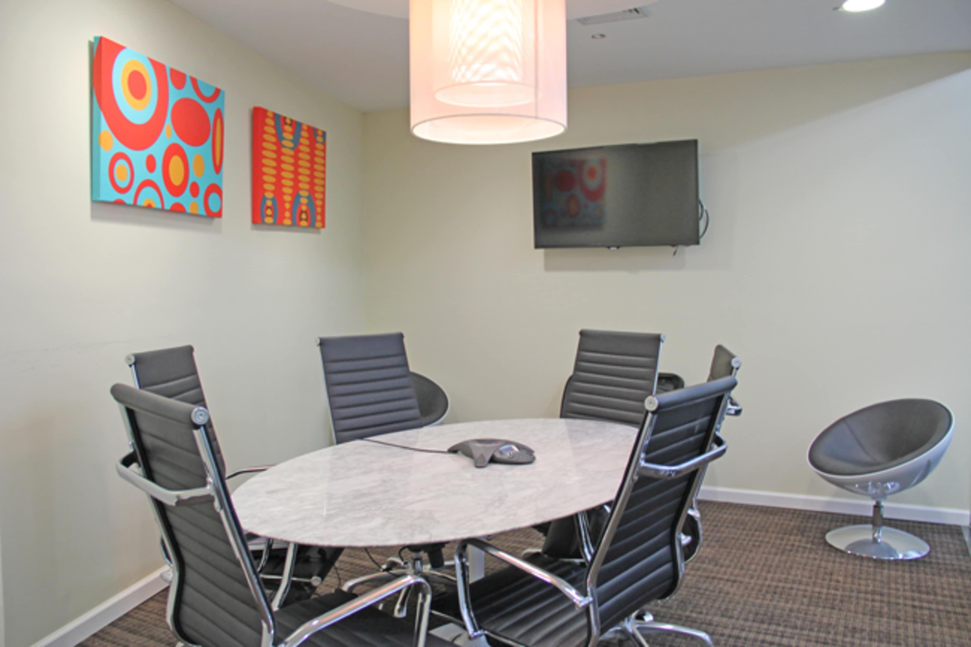 A modern conference room features a round marble table surrounded by six black leather chairs, with colorful abstract art on the walls and a television mounted on one side.
