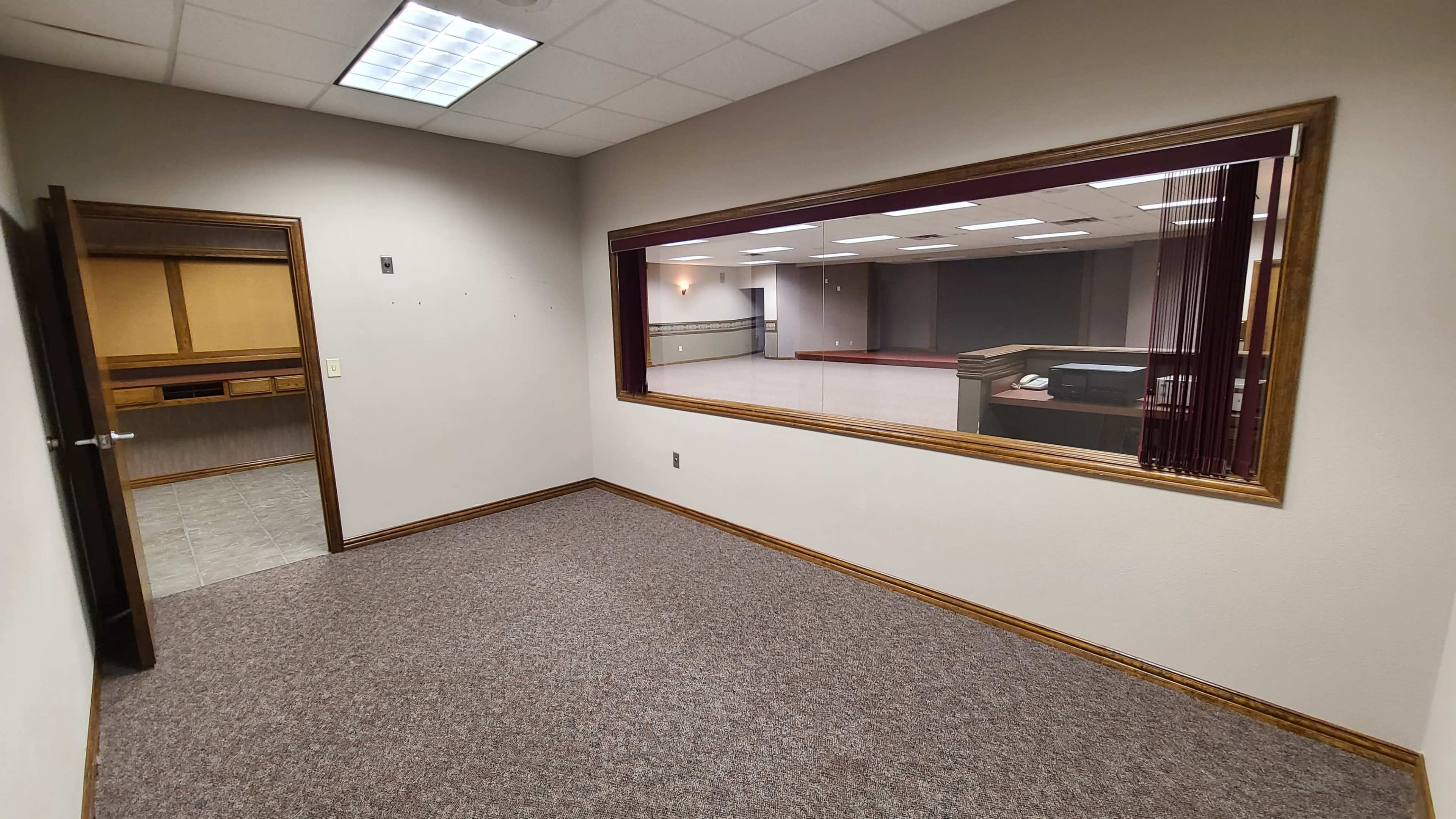 The image shows a vacant office space with gray carpet, beige walls, and a large window facing an adjacent room.