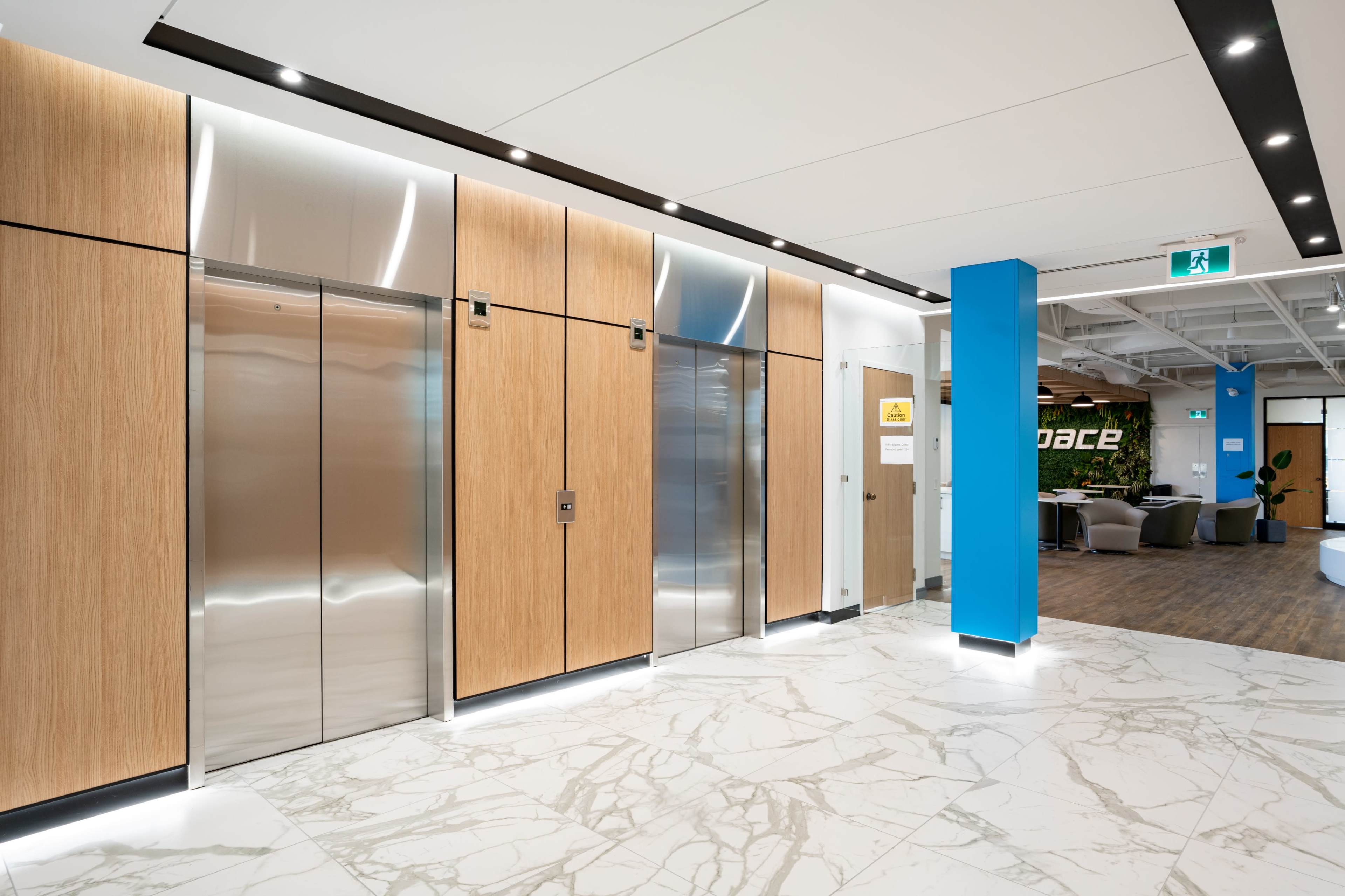 A modern lobby area featuring wooden elevator doors, stainless steel elevators, and a marble floor with a blue accent wall.