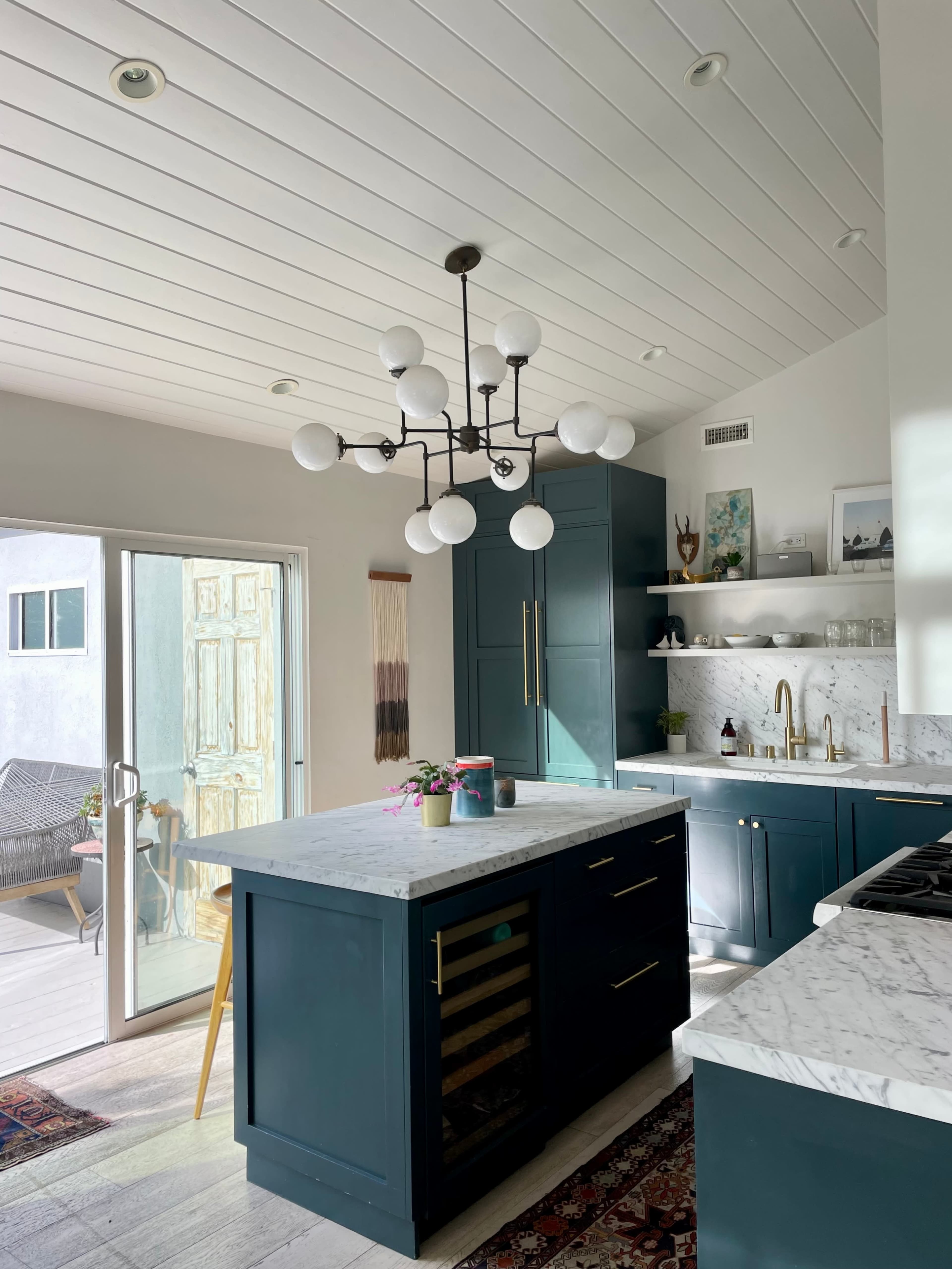 A modern kitchen features blue cabinetry, a marble countertop island with a wine cooler, and a statement chandelier, with large glass doors opening to a patio.