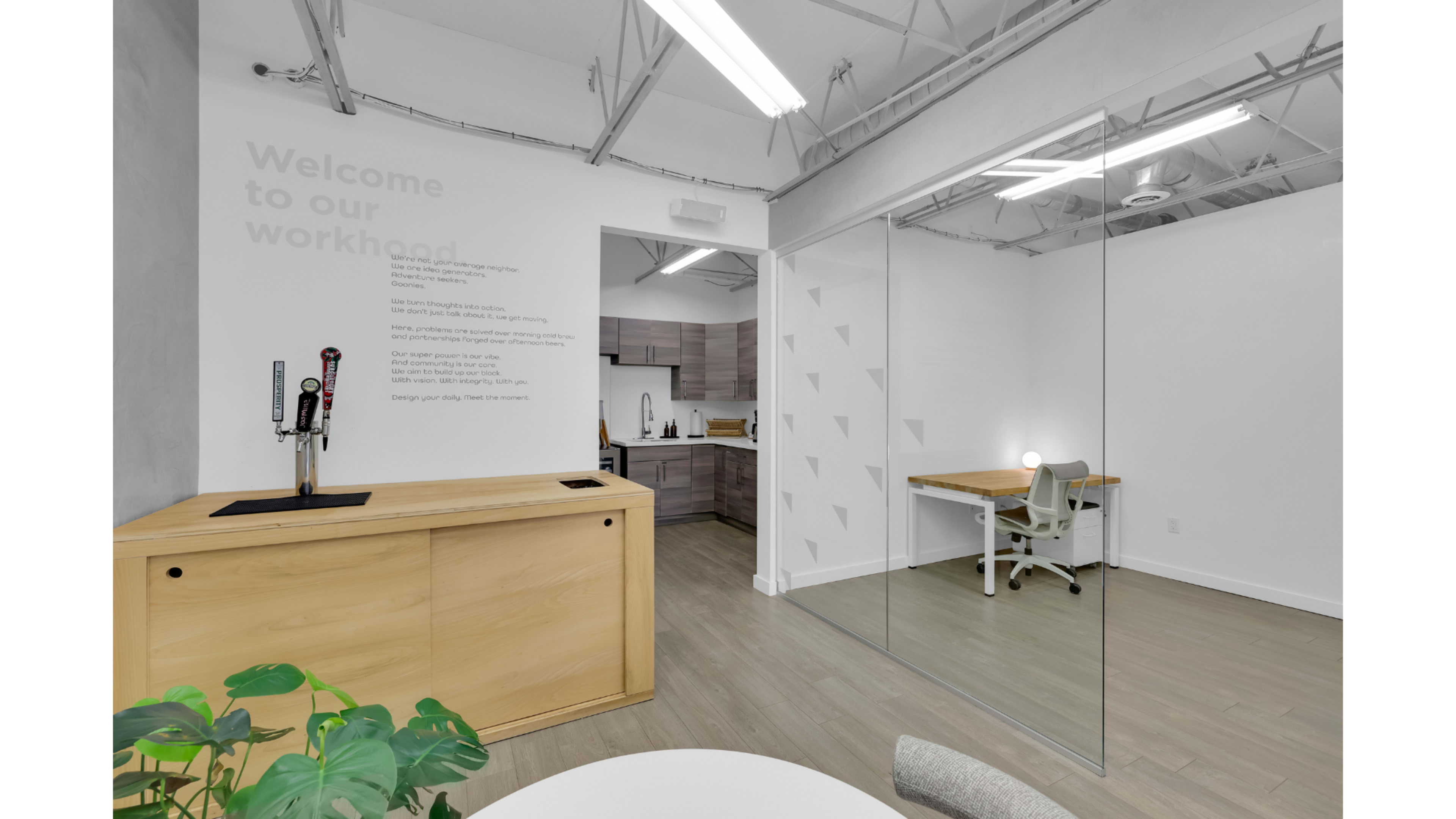 The image shows a modern office space featuring a wooden reception counter, a small kitchenette in the background, and a glass-walled meeting area with a desk and chair.