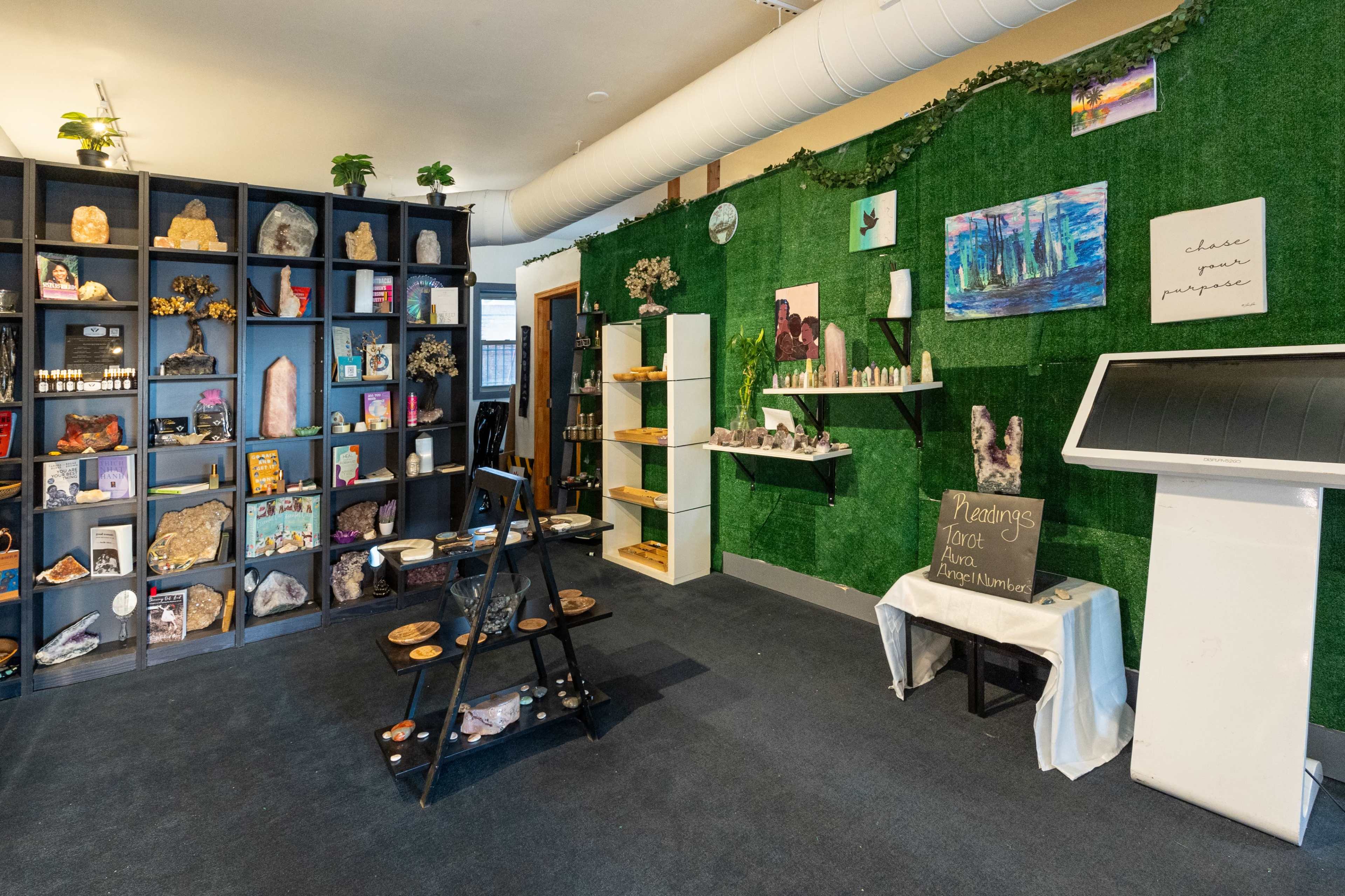 A bright, organized space featuring shelves with crystals and spiritual items, a small display table in the center, and green wall decor.