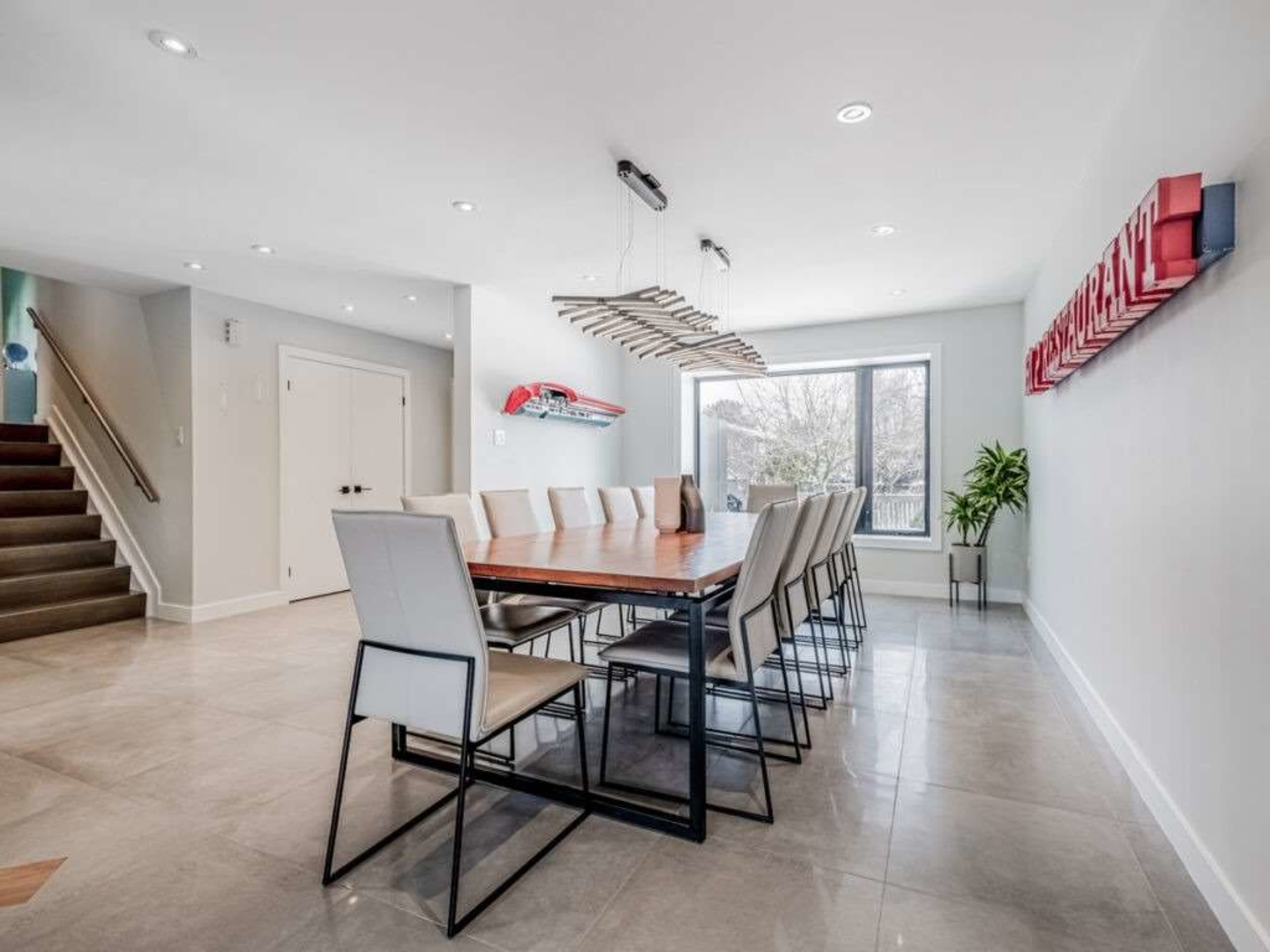 The image shows a modern dining area with a long wooden table surrounded by light-colored chairs, large windows, and minimalistic decor.