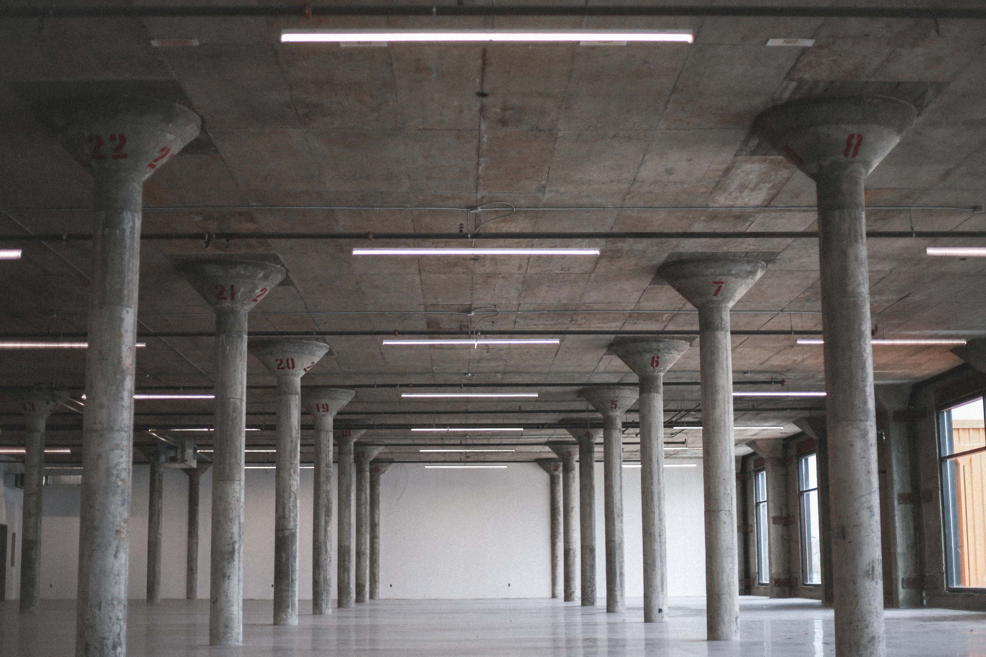 The image shows a spacious, empty industrial interior with exposed concrete columns and overhead lighting.