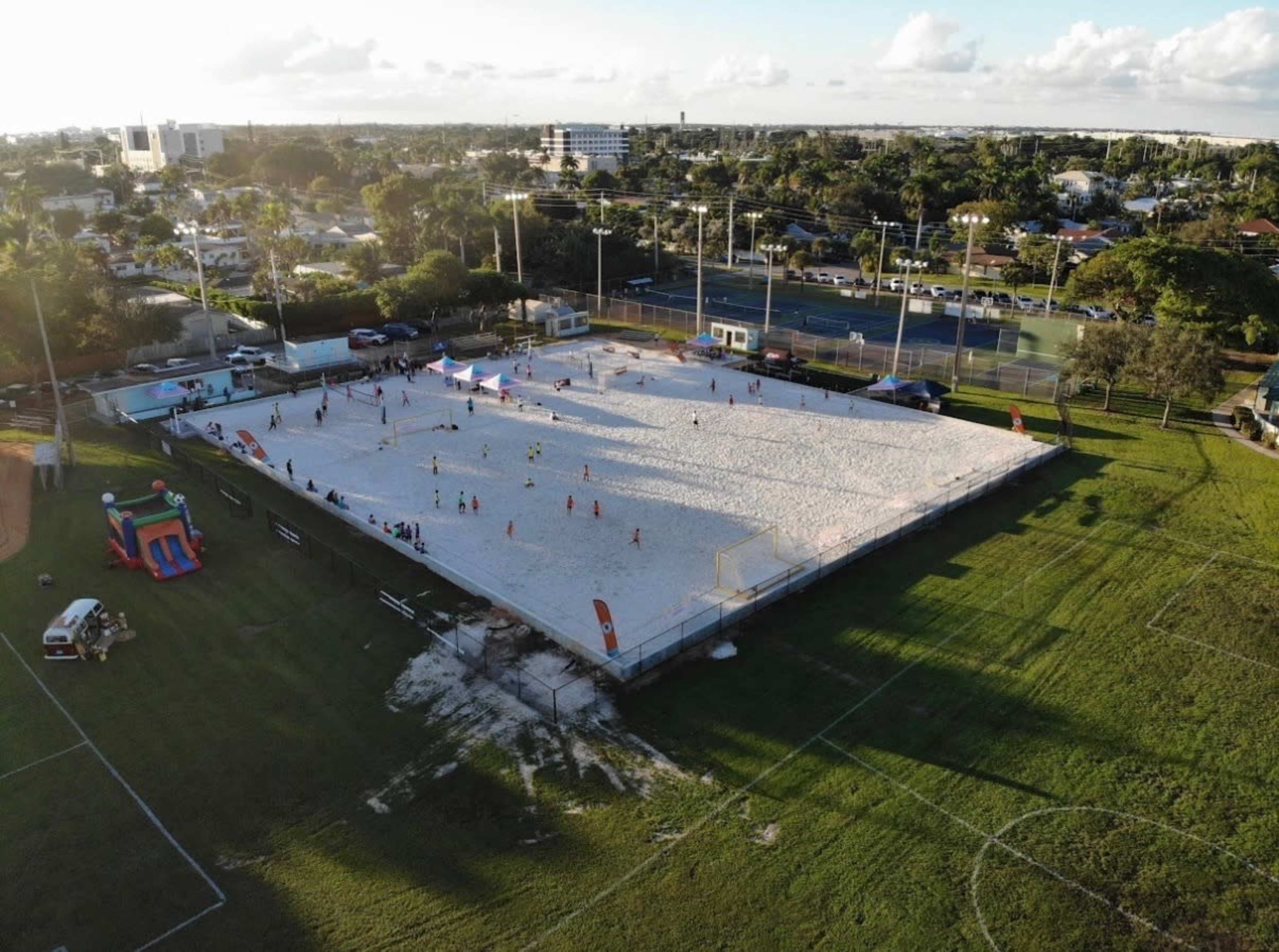 The image shows a sandy beach volleyball court with several players engaged in a game, surrounded by green grass and facilities, including playground equipment and sports fields in the backdrop.