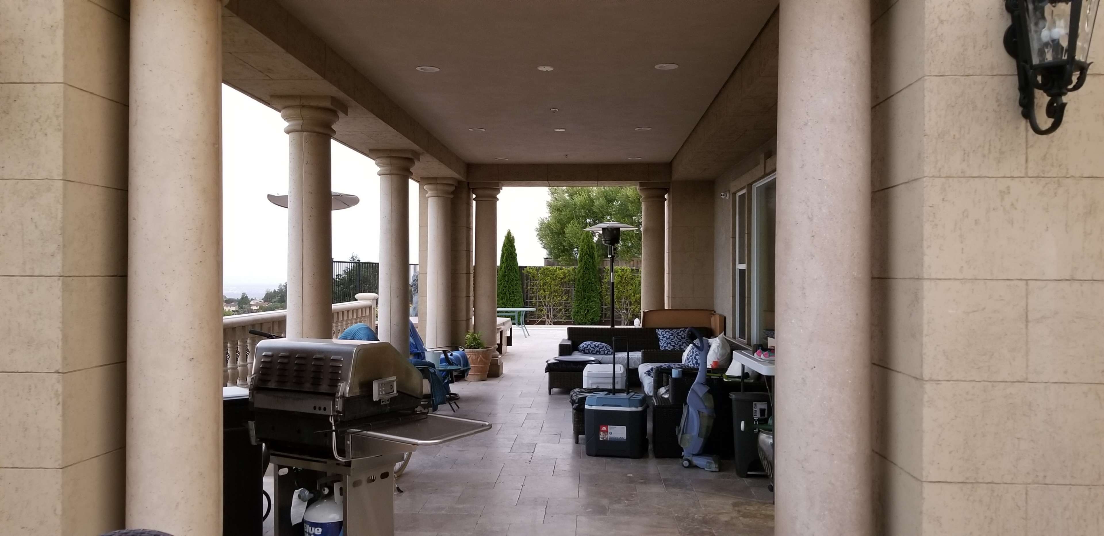 A covered patio with columns features a barbecue grill and various boxes and furniture positioned along the edges.