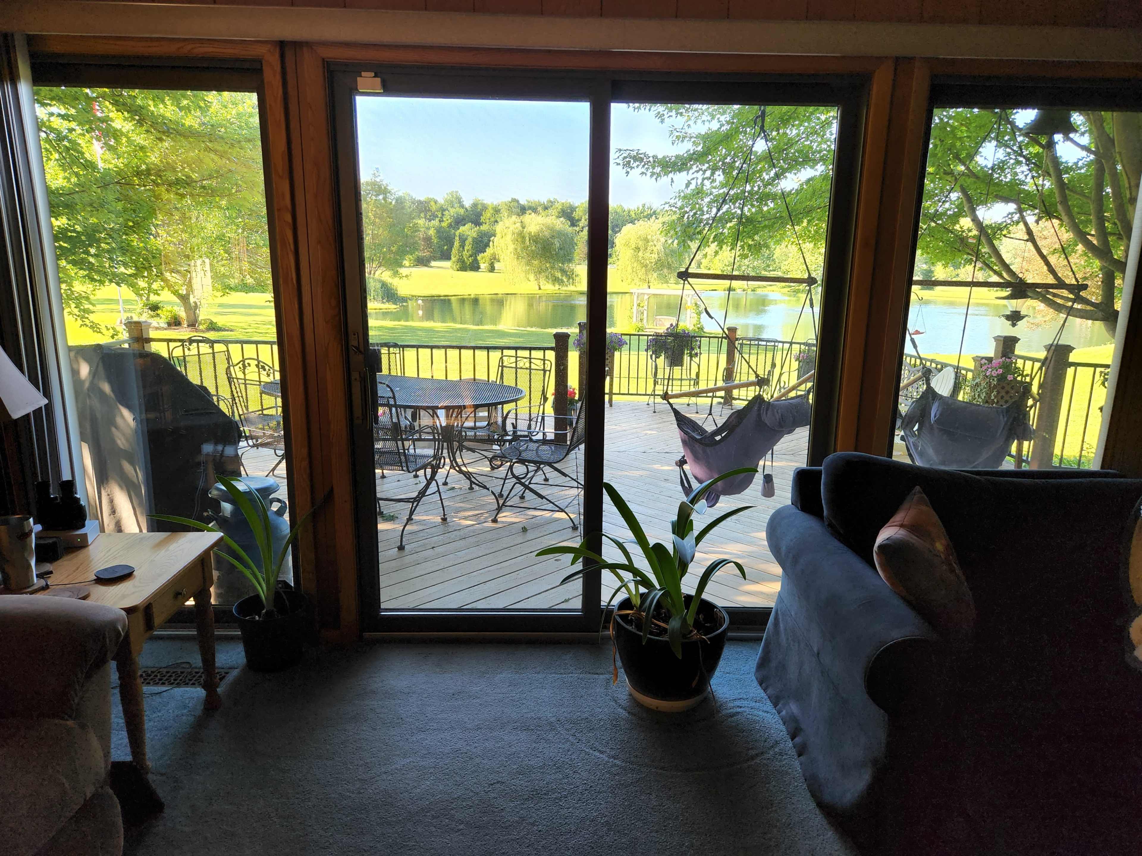 A view from inside a home through sliding glass doors, revealing a deck with chairs overlooking a lake surrounded by greenery.