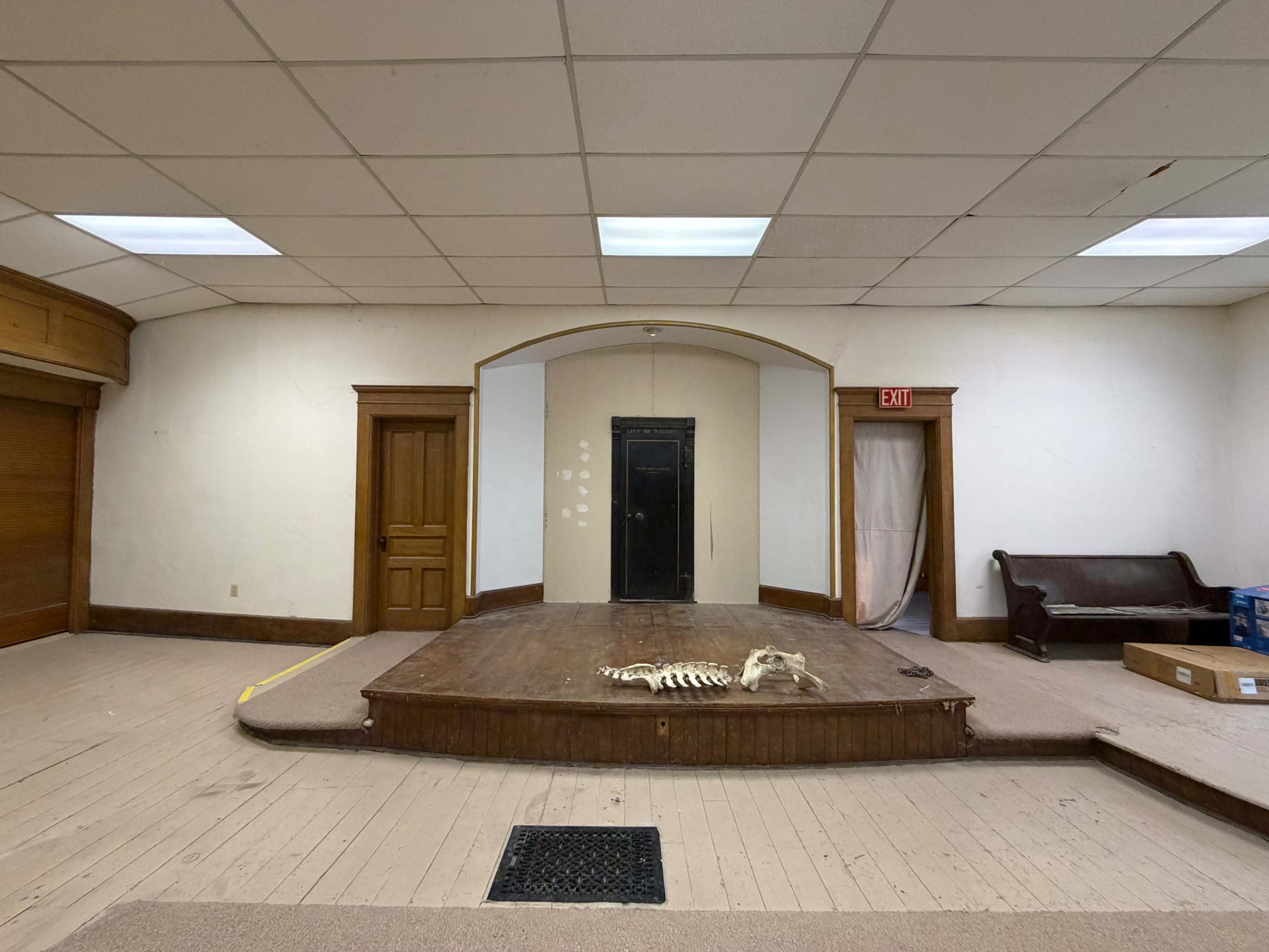The image shows a vacant stage with a wooden platform, a skeleton lying on the floor, and a backdrop featuring a black door in a plain room with white walls.