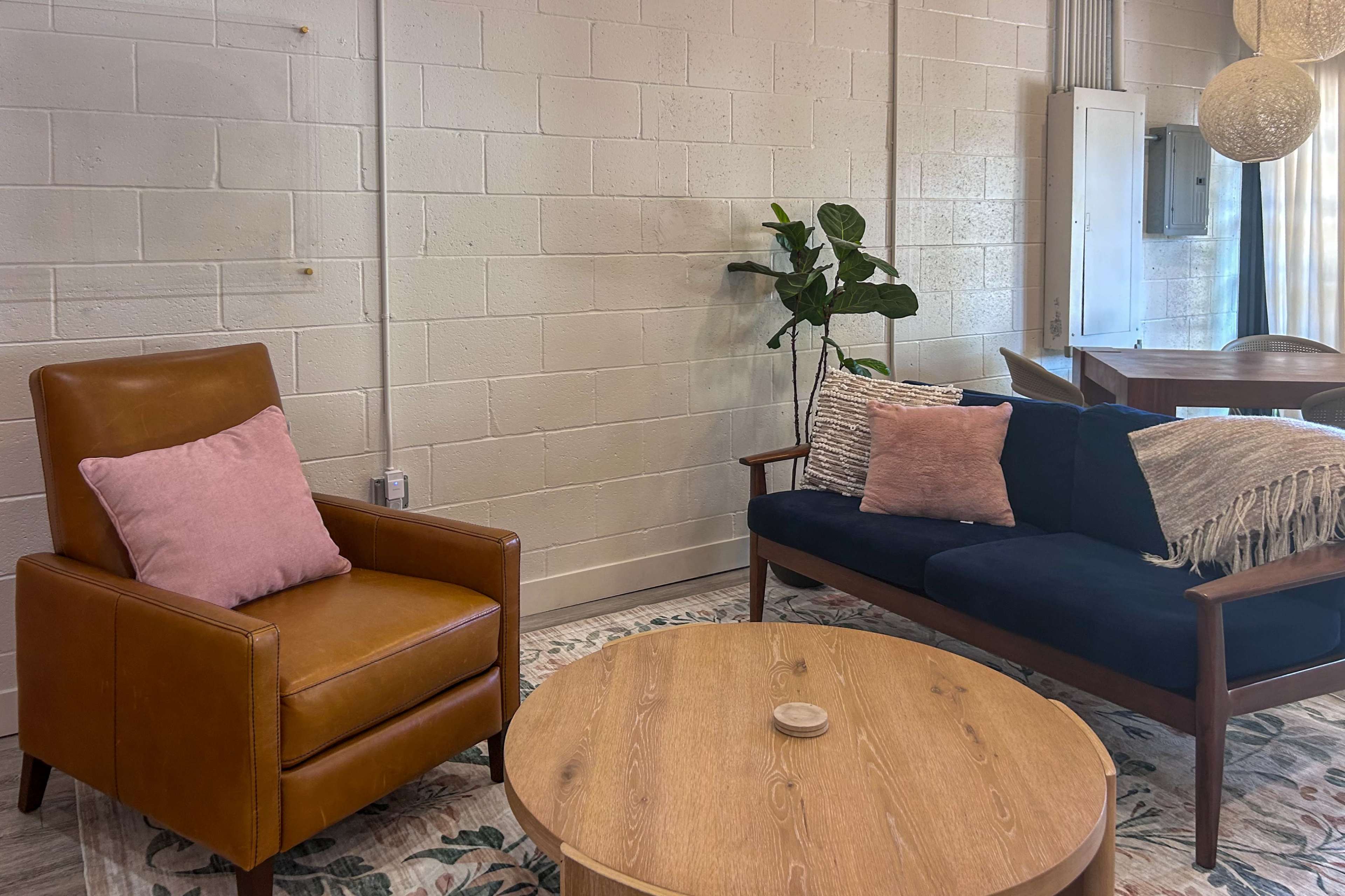 The image shows a cozy living area with a brown leather armchair, a dark blue sofa with pink accent pillows, a round wooden coffee table, and a potted plant in a corner.
