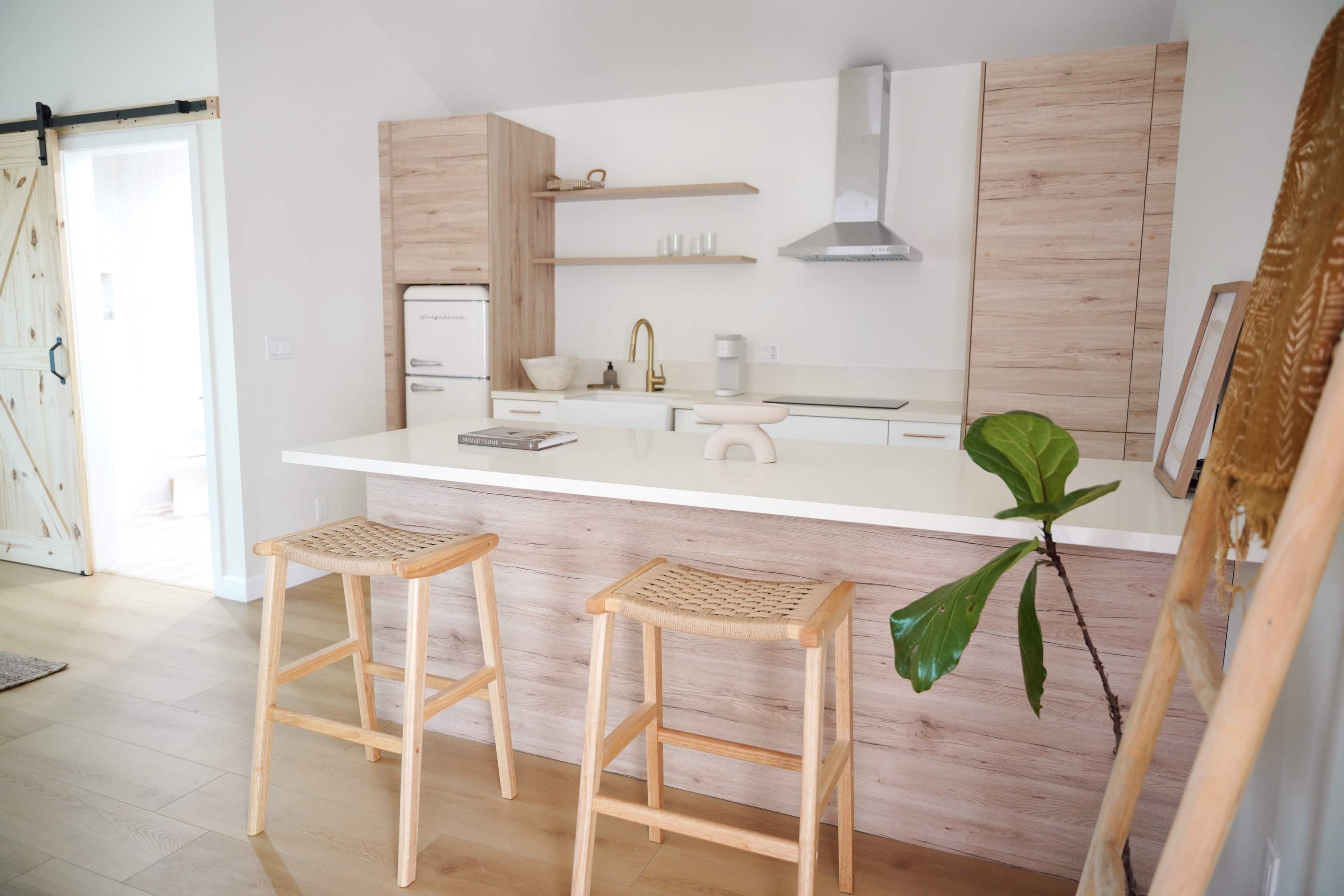 A modern kitchen features light wood cabinetry, a white countertop, and two wicker bar stools.