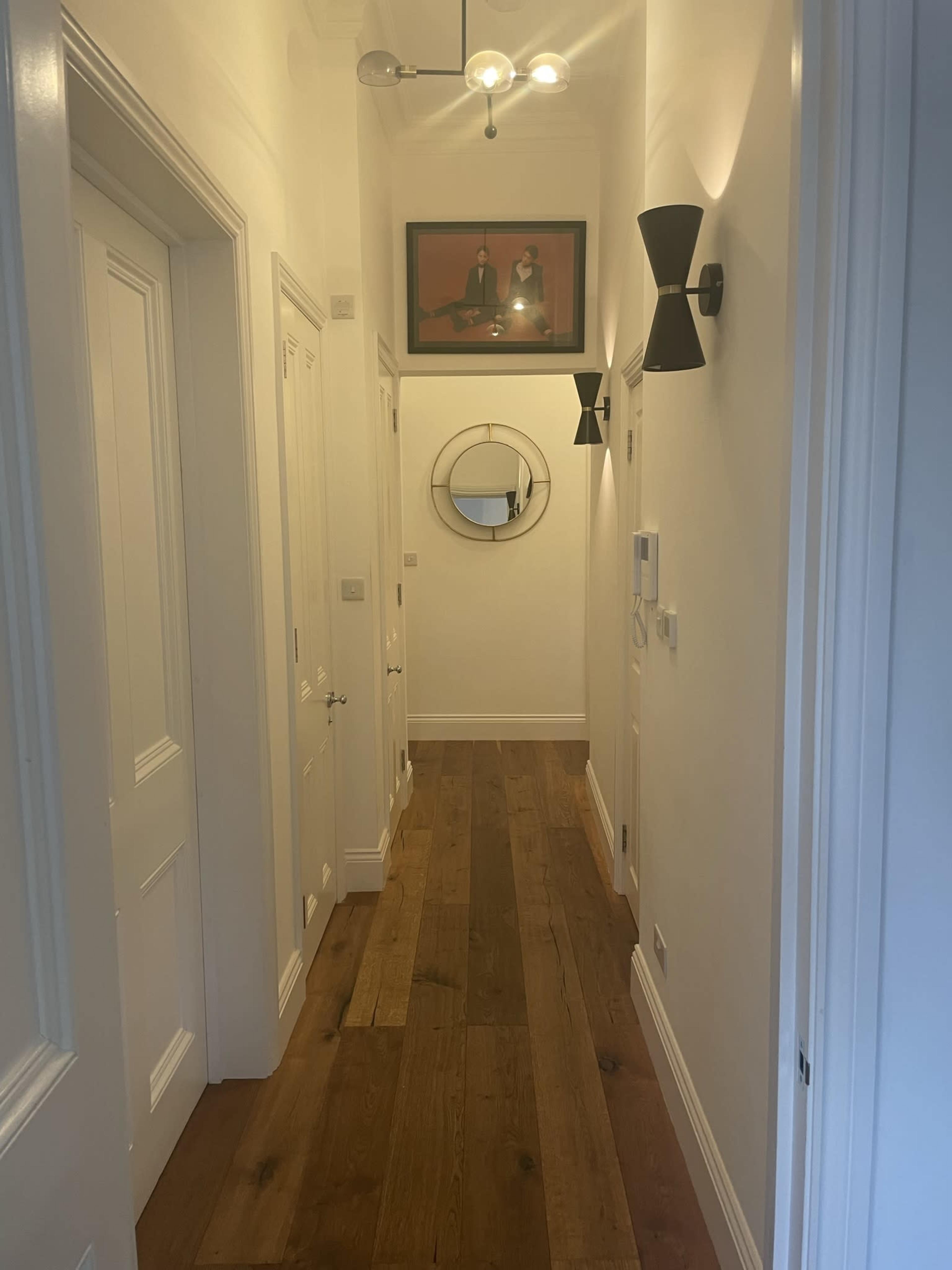 A narrow hallway features white walls, wooden flooring, and framed art along with a round mirror and modern wall sconces.