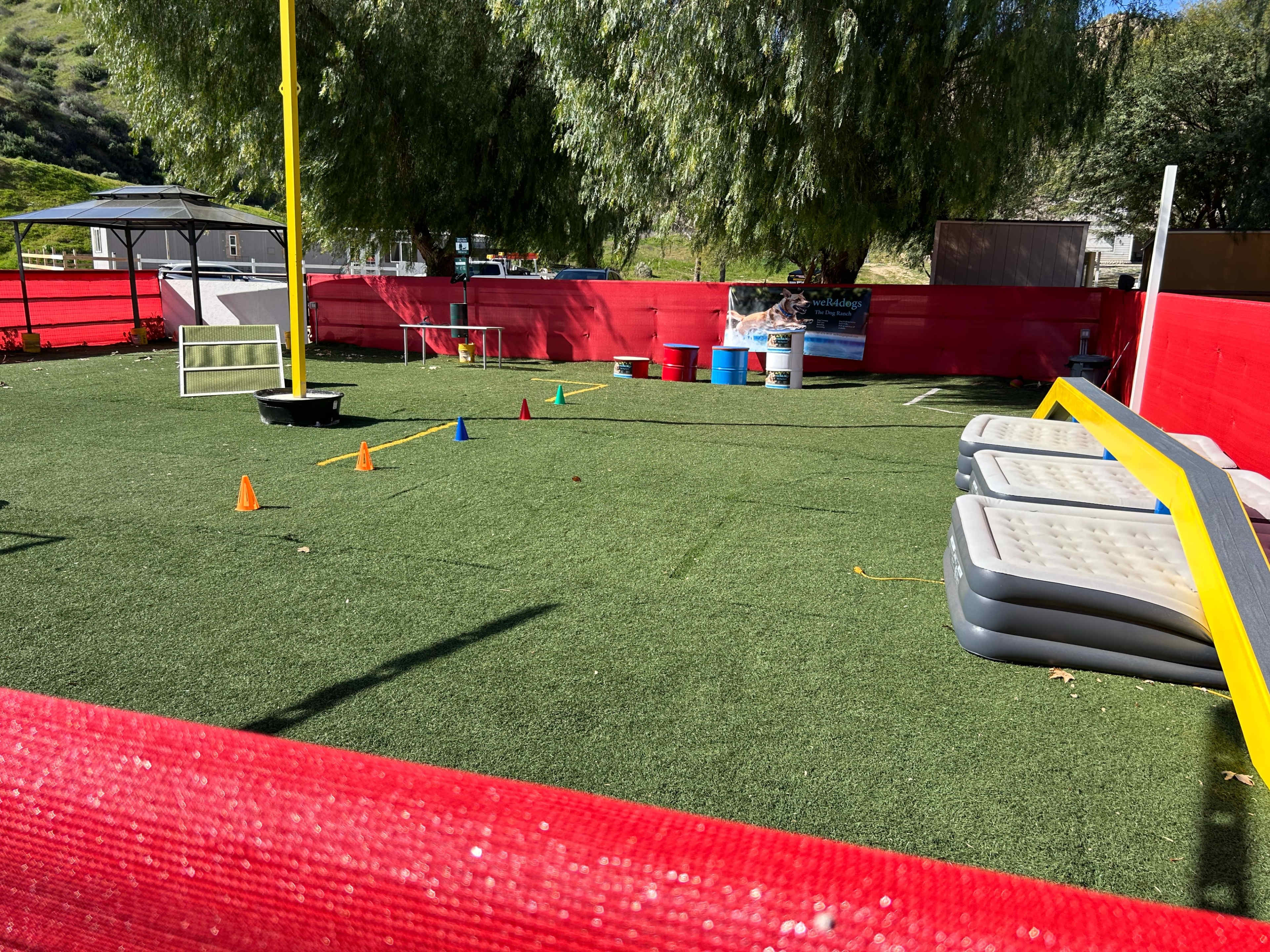 The image shows an outdoor training area with artificial turf, featuring cones arranged on the ground, a goalpost, and some large inflatable mattresses along the side.