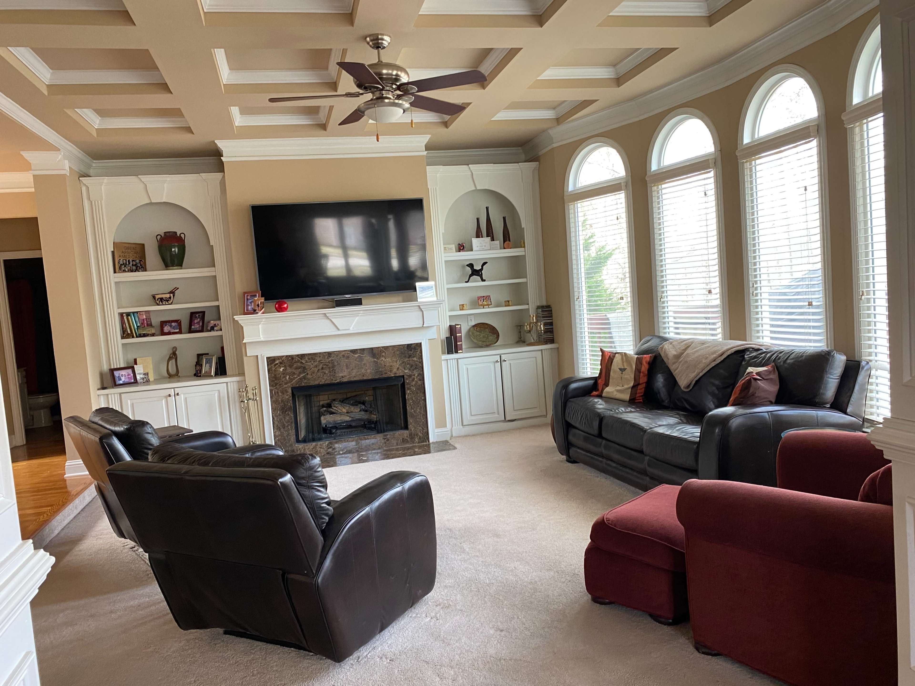 A living room features a coffered ceiling, a television mounted above a fireplace, and three seating arrangements, including a red chair, in front of large windows.