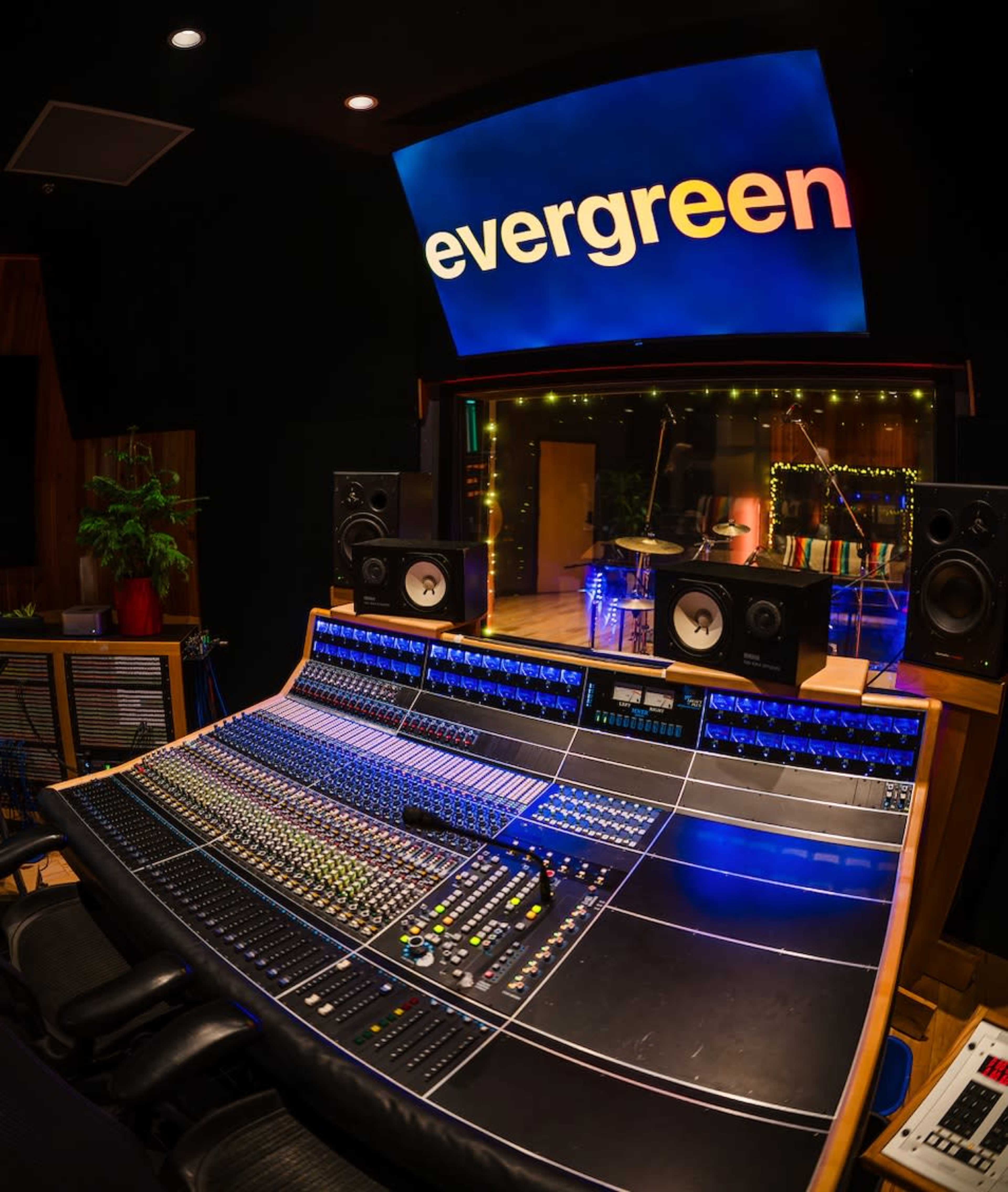 The image shows a music recording studio featuring a large mixing console, monitors, and a brightly lit area with instruments in the background.