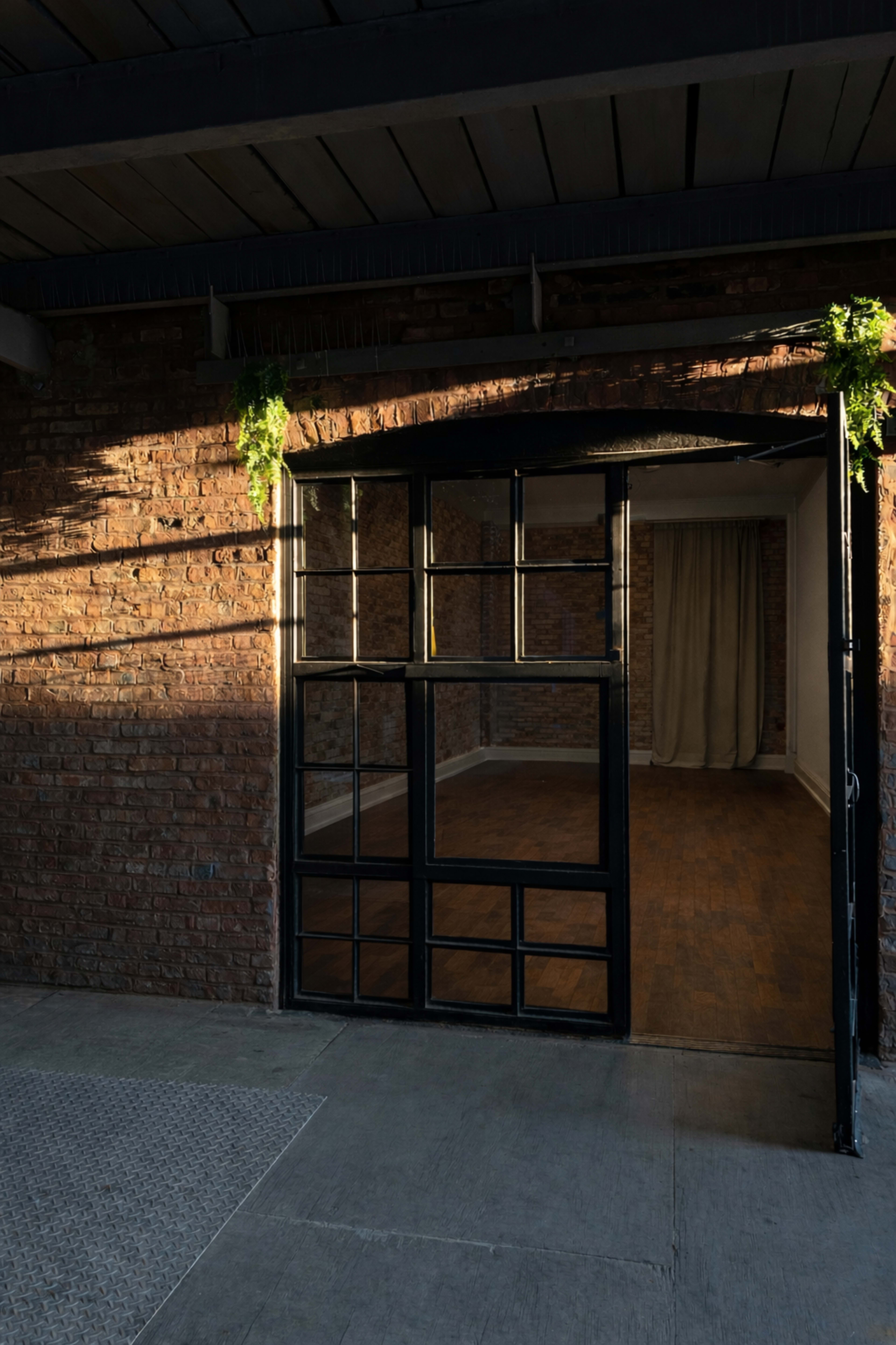 A black-framed glass door opens to reveal a well-lit, empty room with wooden flooring and a curtain in the background.