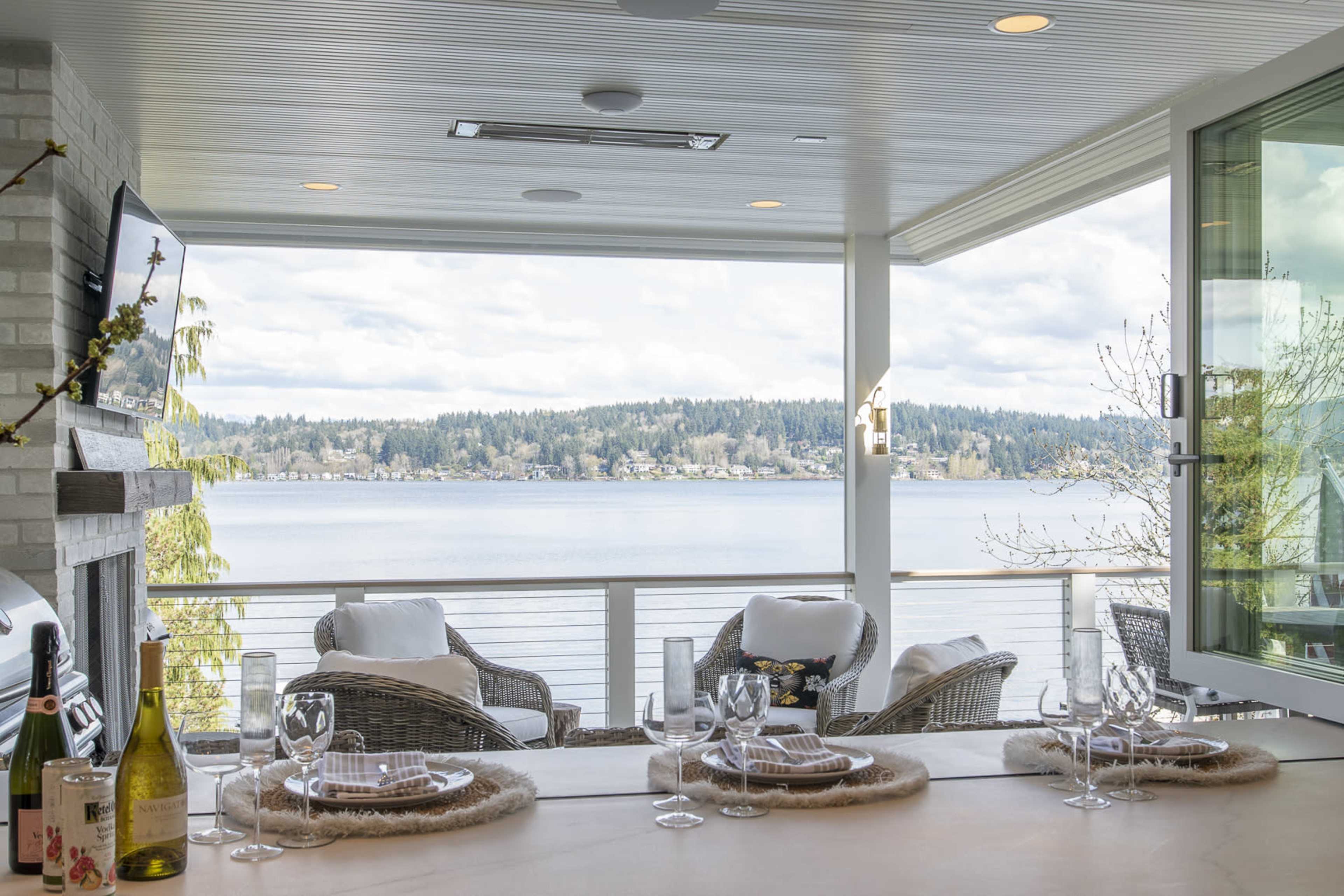 A scenic view of a lakeside terrace features a dining setup with wine glasses and a bottle, overlooking calm waters and a forested shoreline.