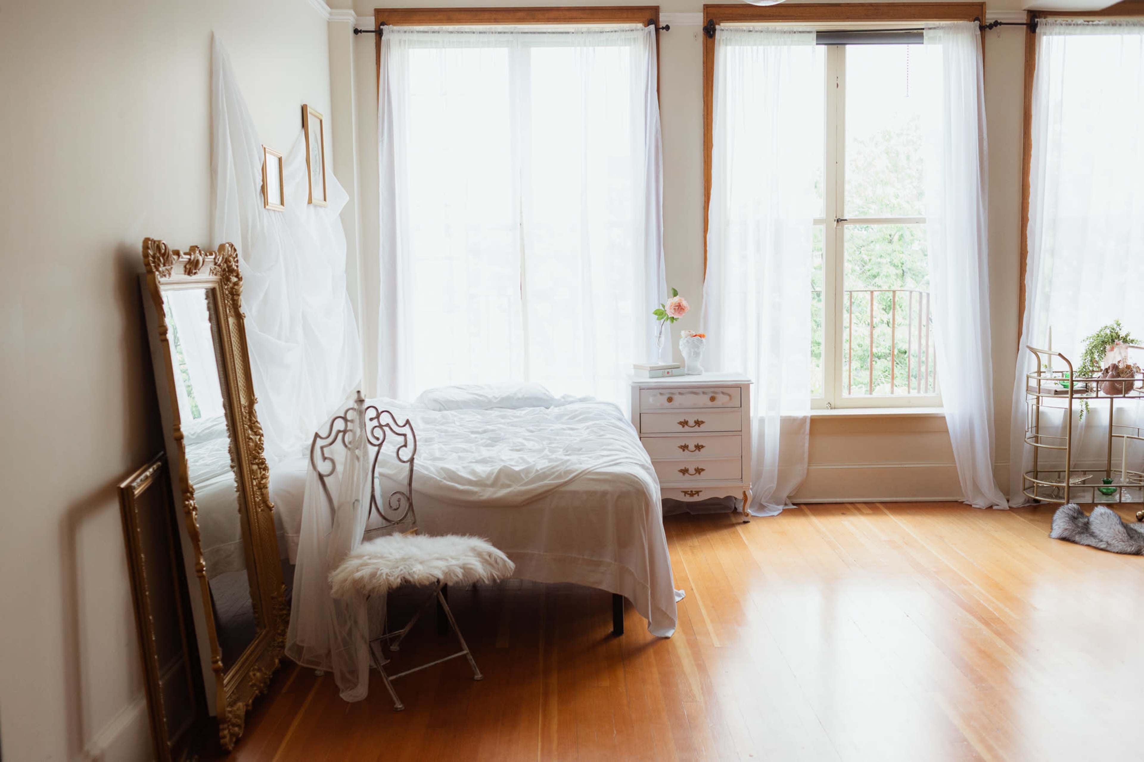 A softly lit bedroom features a bed draped in white fabric, a large ornate mirror, and a small nightstand beside a window with sheer curtains.
