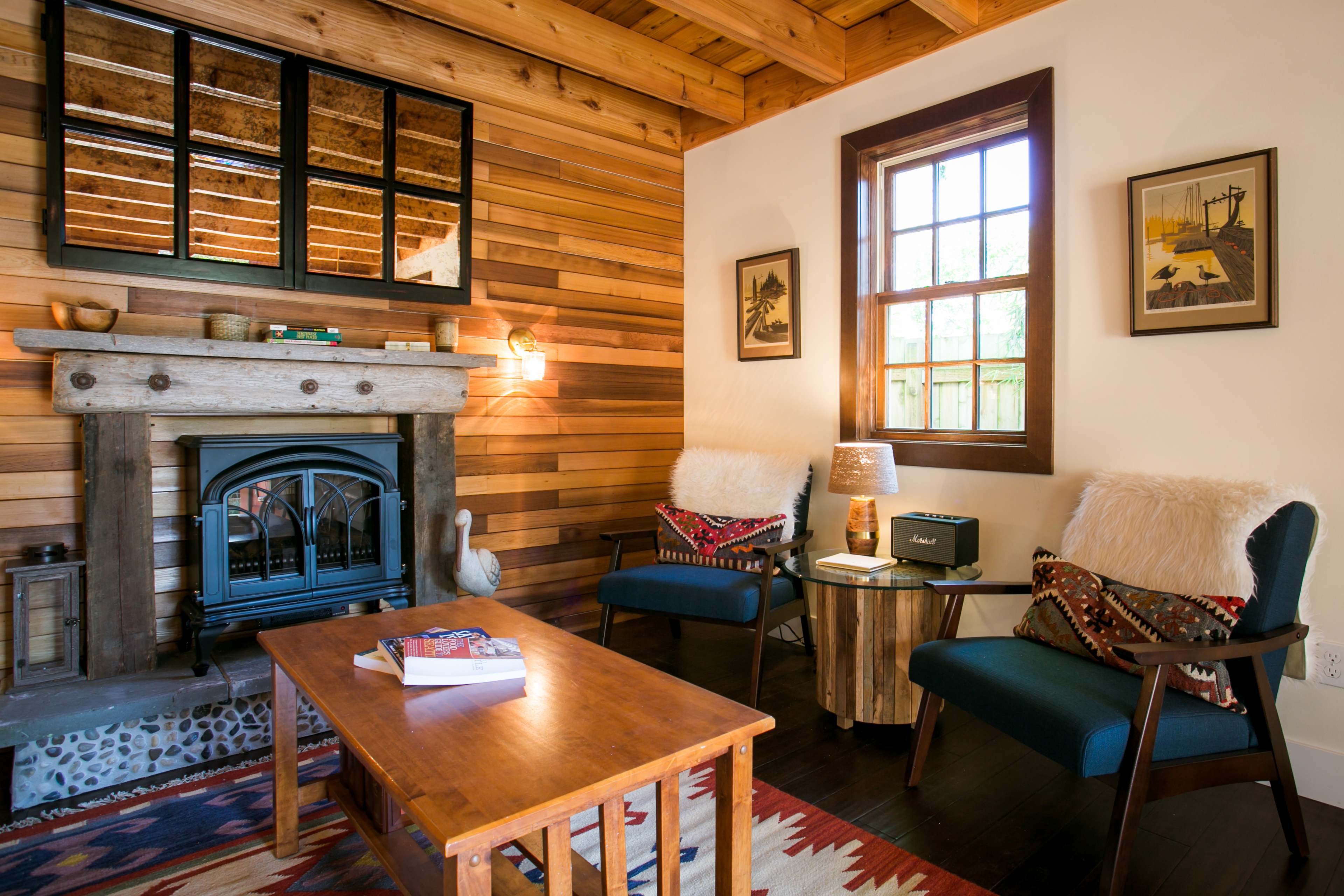 A cozy living room featuring a wooden ceiling, a stone fireplace with a mantel, two chairs with decorative throw pillows, and a wooden coffee table.