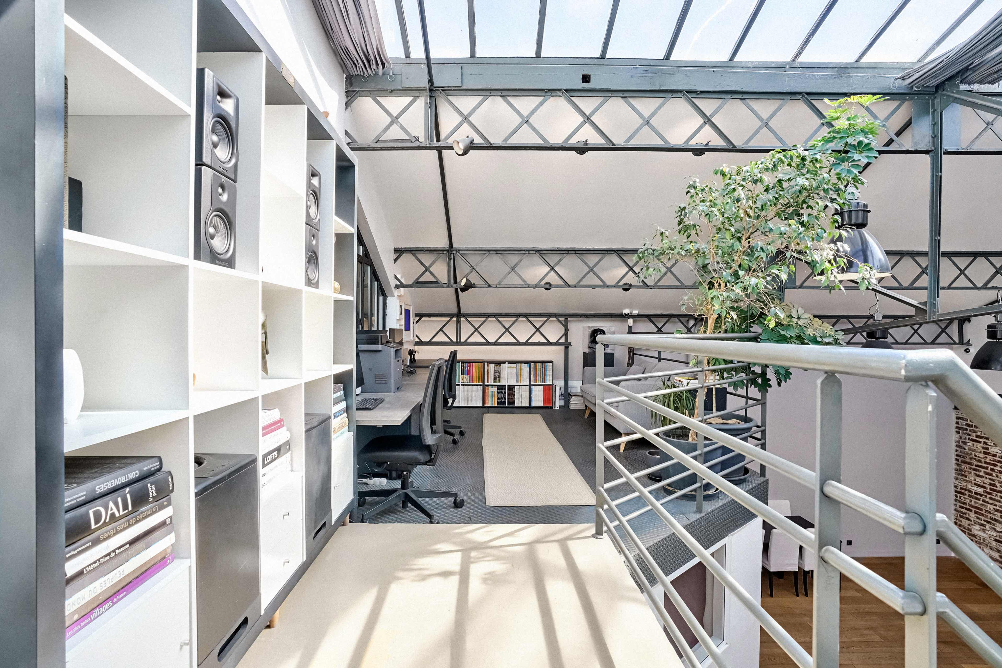 The image shows a loft-style workspace with a railing overlooking a lower level, featuring white shelving, a desk with speakers, and a potted plant.