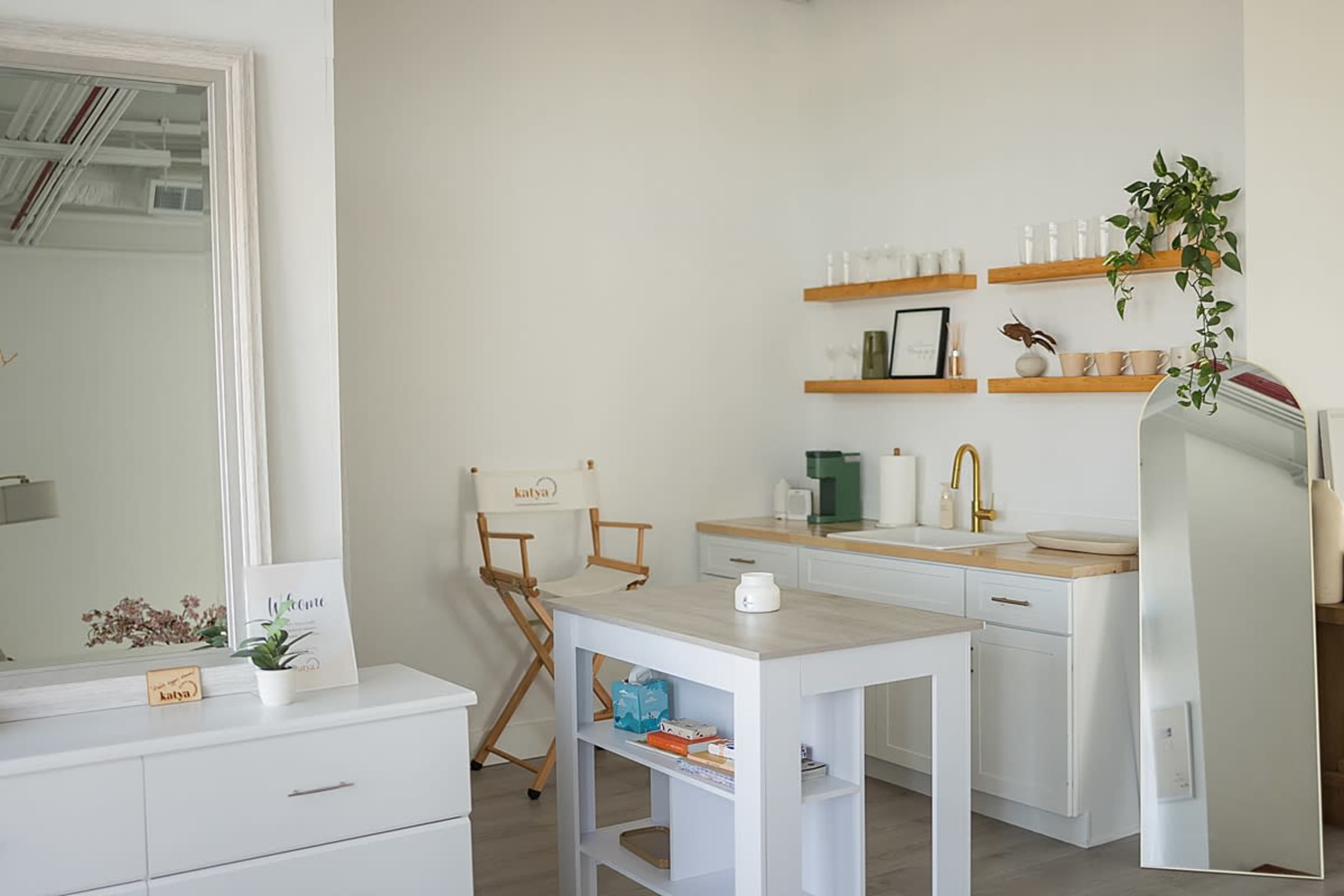 The image shows a bright, minimalist workspace featuring a small kitchen area with wooden shelves and a reflection in a mirror.