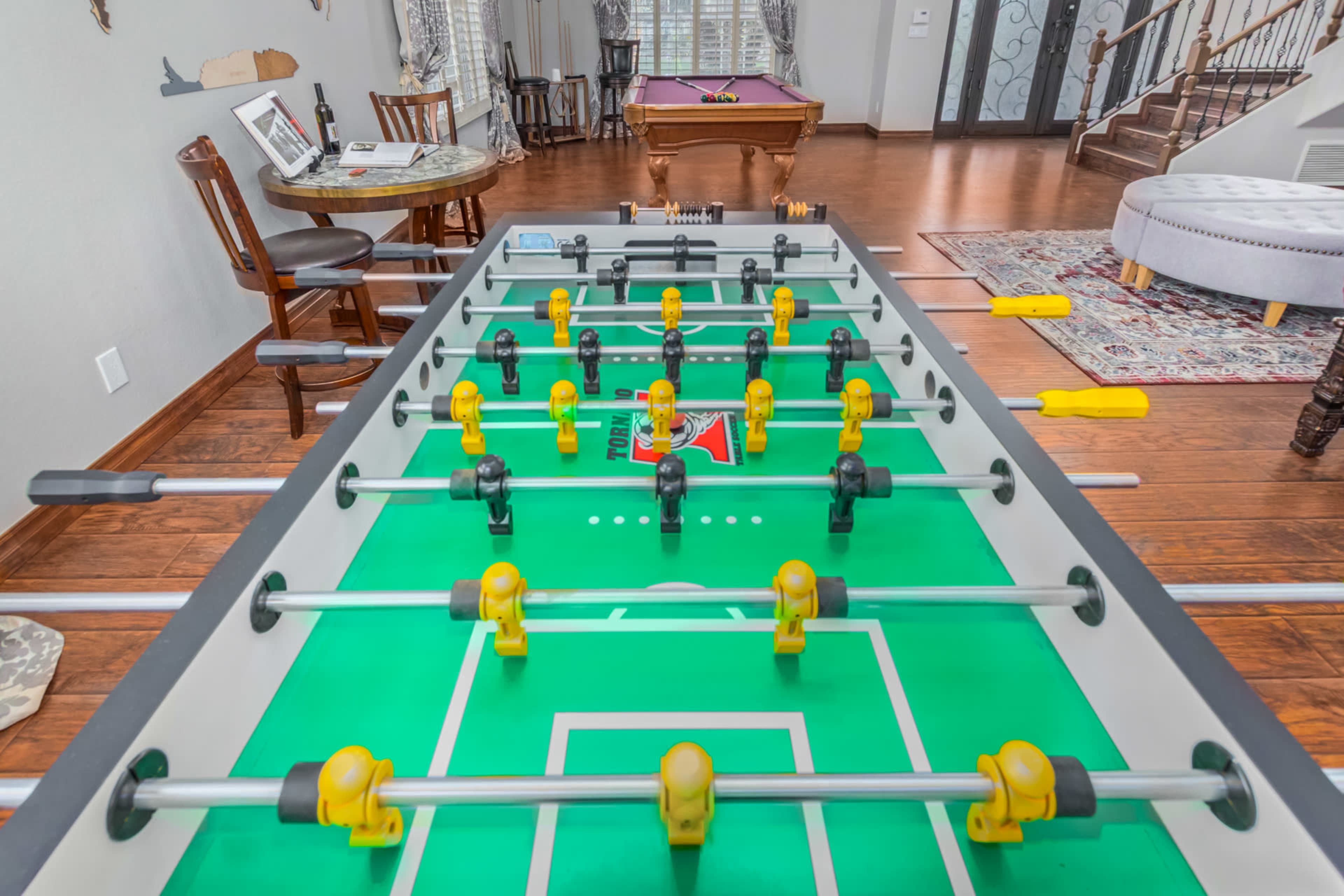 A foosball table is set up in a living room with a pool table in the background and various furniture around.