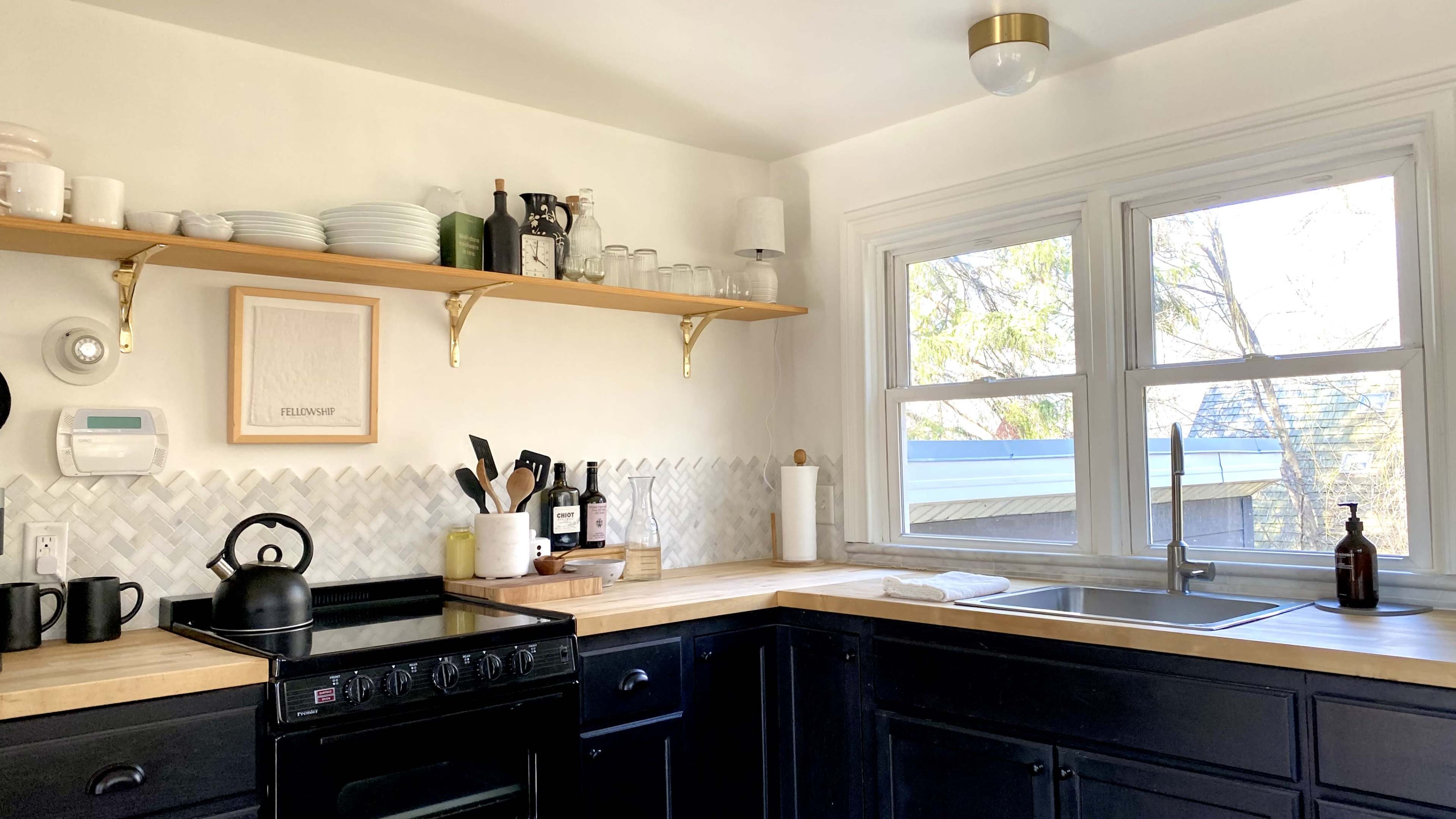 A modern kitchen features black cabinetry, a wooden countertop with utensils, shelves displaying dishware, and large windows providing natural light.