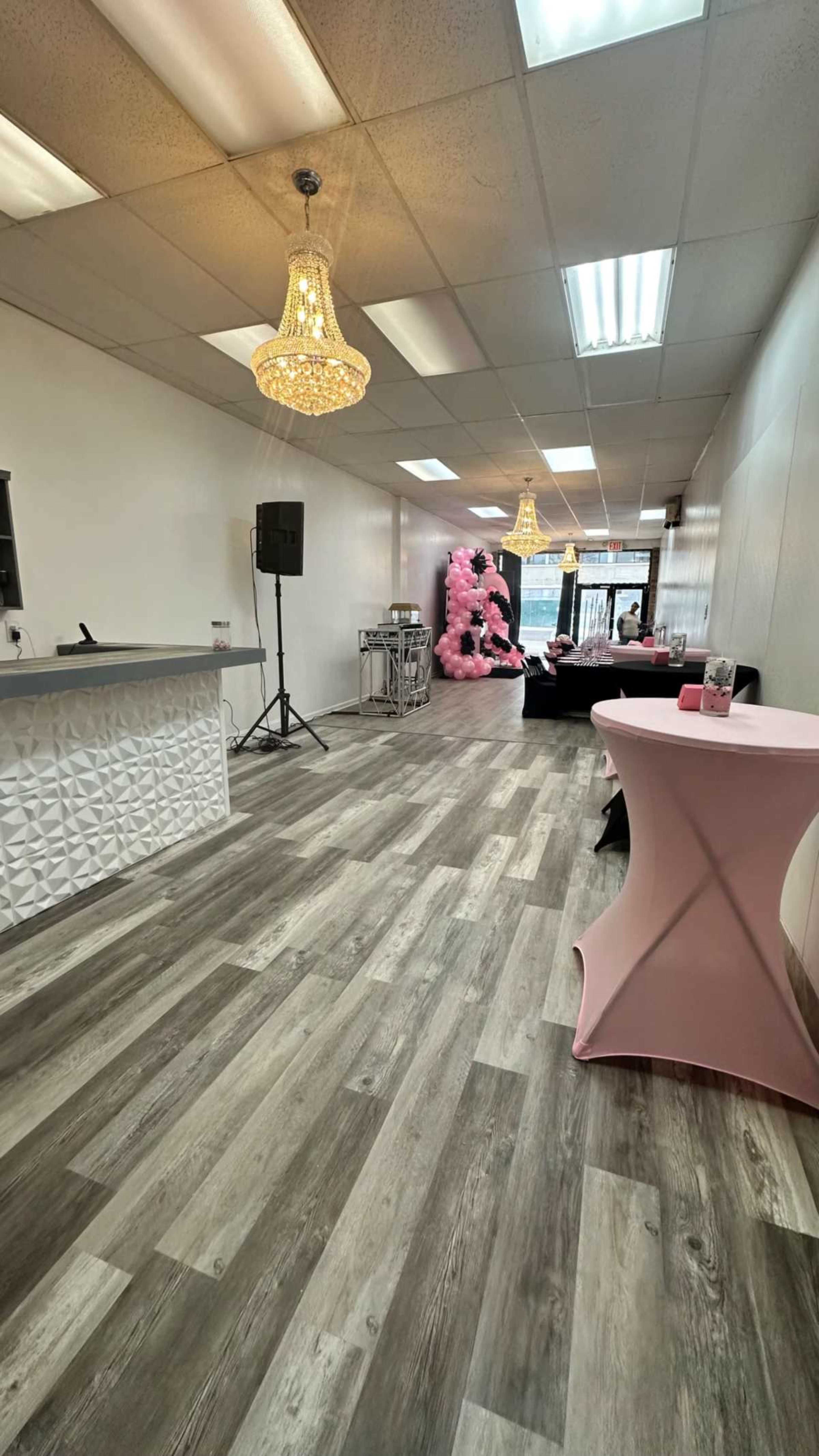 The image shows a spacious interior of a venue featuring a modern stone accent wall, a bar area, pink decor, and two decorative chandeliers hanging from the ceiling.