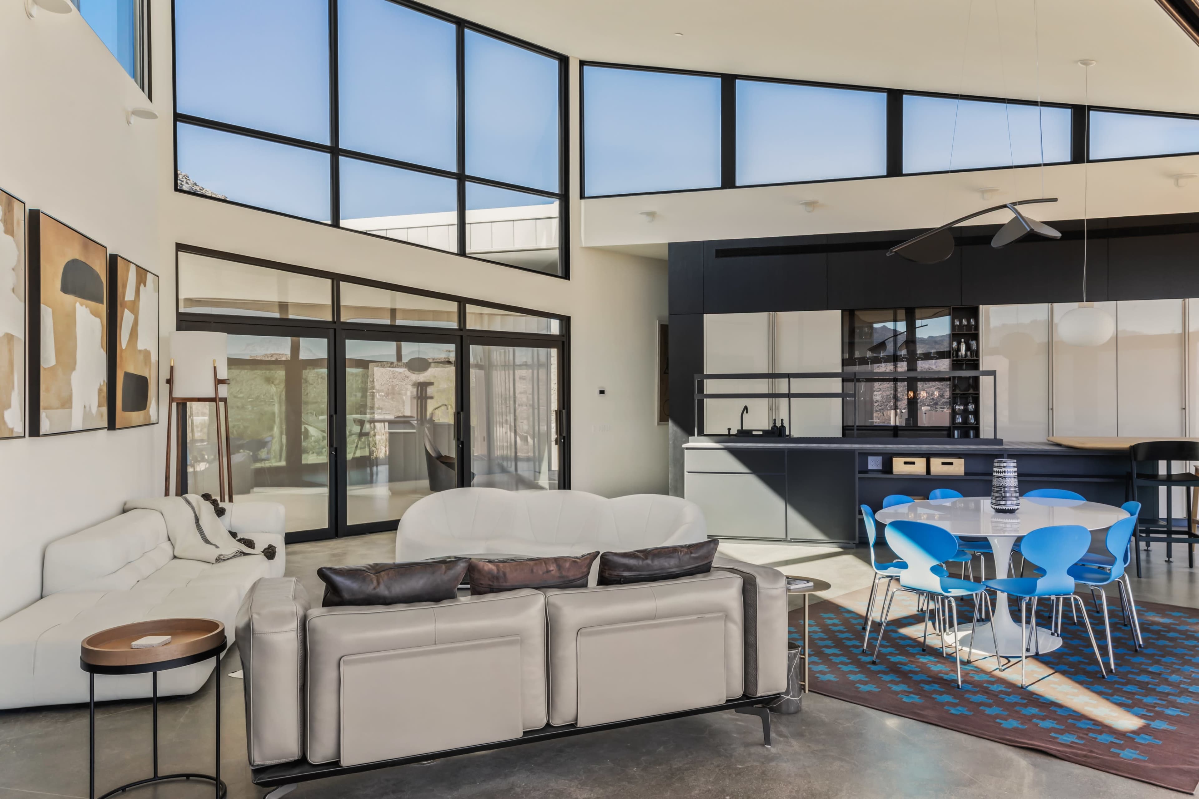 The image shows a modern living space with large windows, sleek furniture, and a dining area featuring a round table and blue chairs.