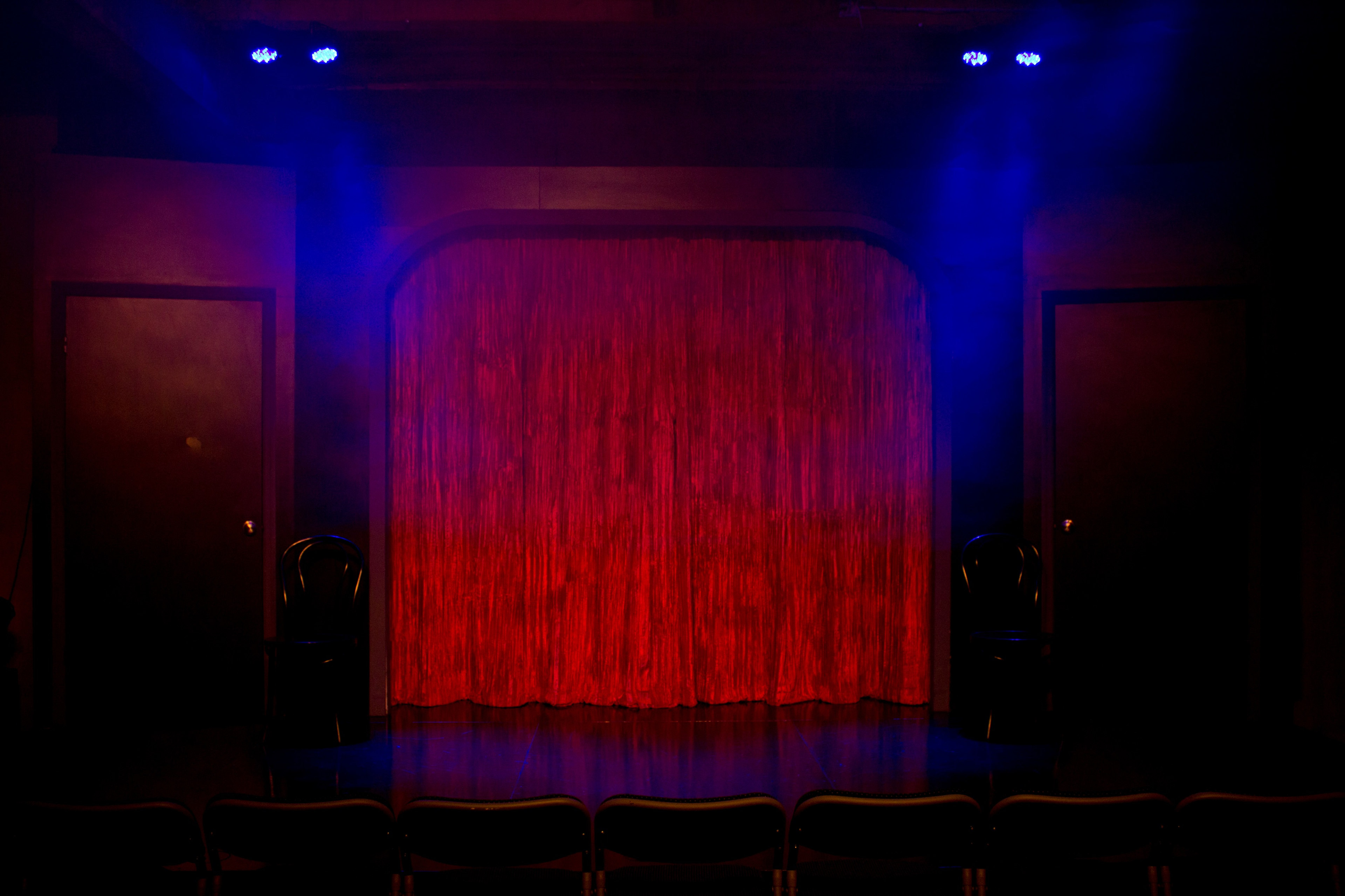 A dimly lit stage with a red curtain and two chairs positioned on either side, ready for a performance.
