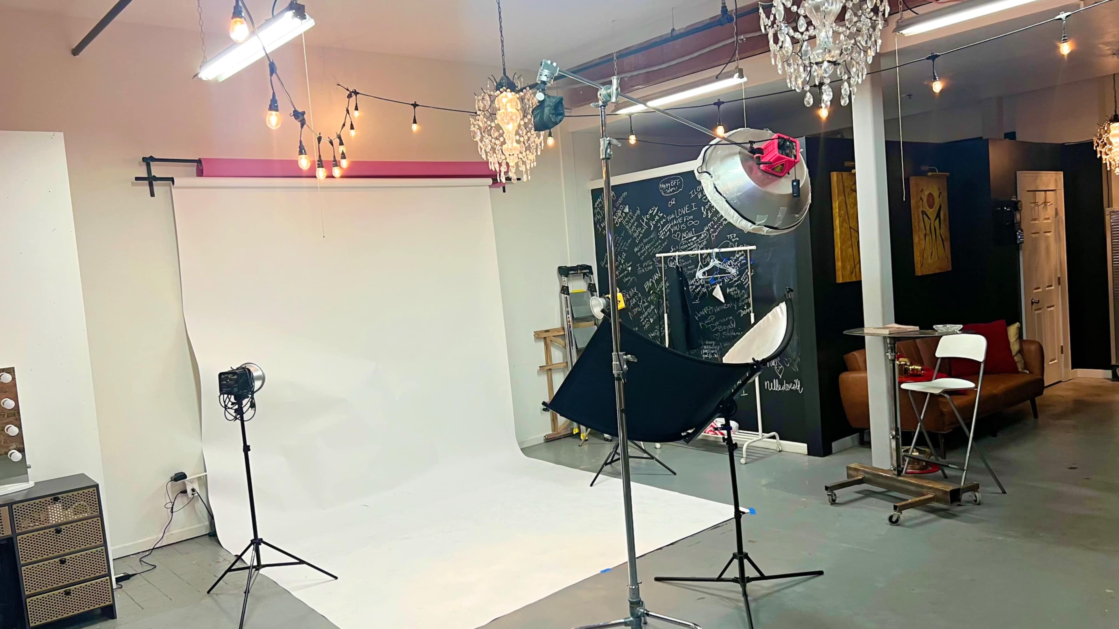 The image shows a photography studio equipped with a white backdrop, various lighting setups, and decorative chandeliers, along with a seating area.