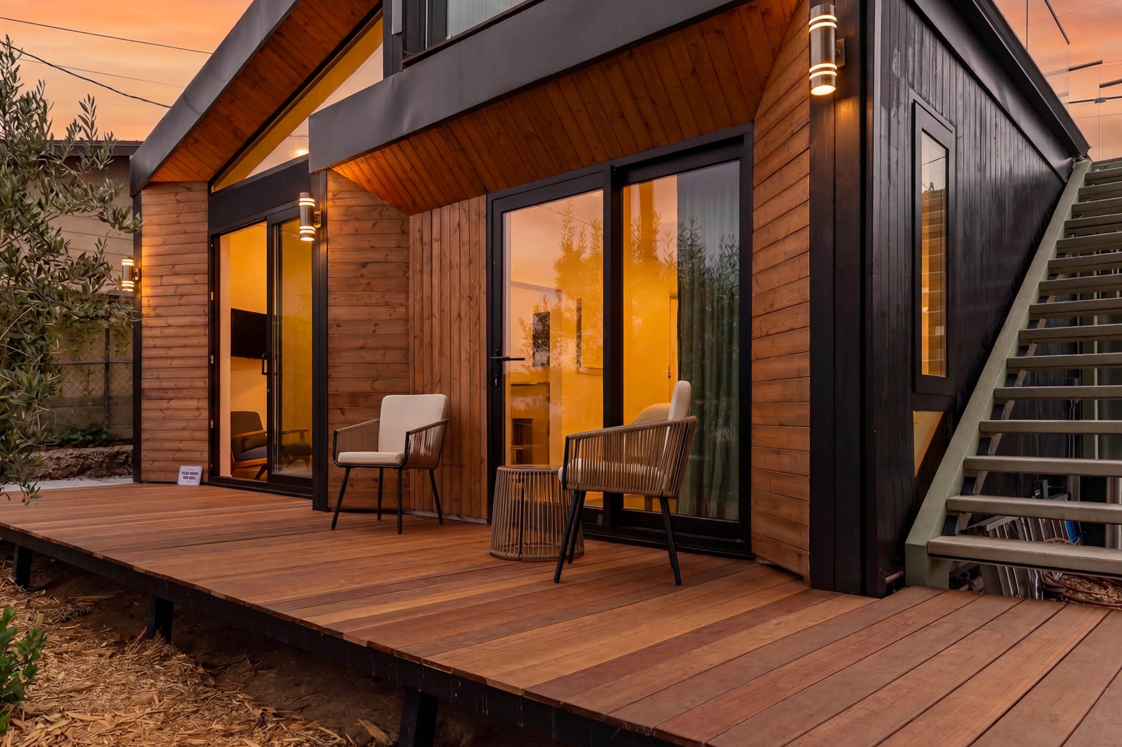 A modern house features large glass doors leading to a wooden deck with two chairs, surrounded by greenery, under a colorful sky at dusk.