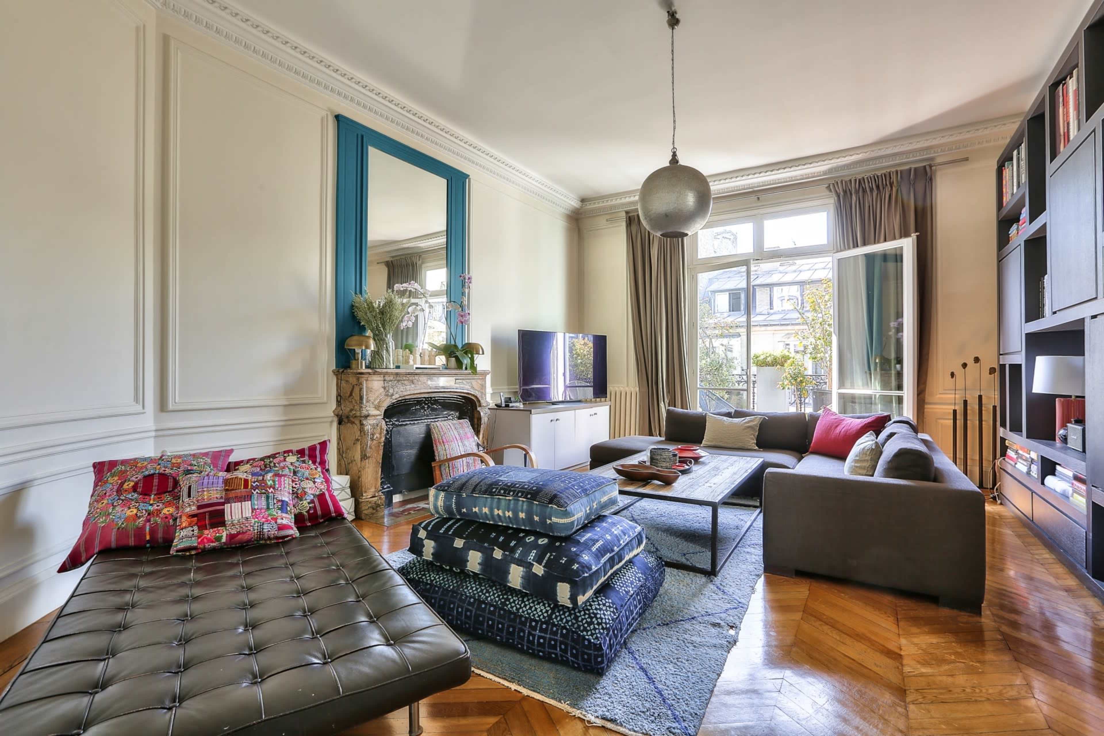 The living room features a mix of patterned cushions on a dark sofa, a glass coffee table, and a large window that opens to a garden.