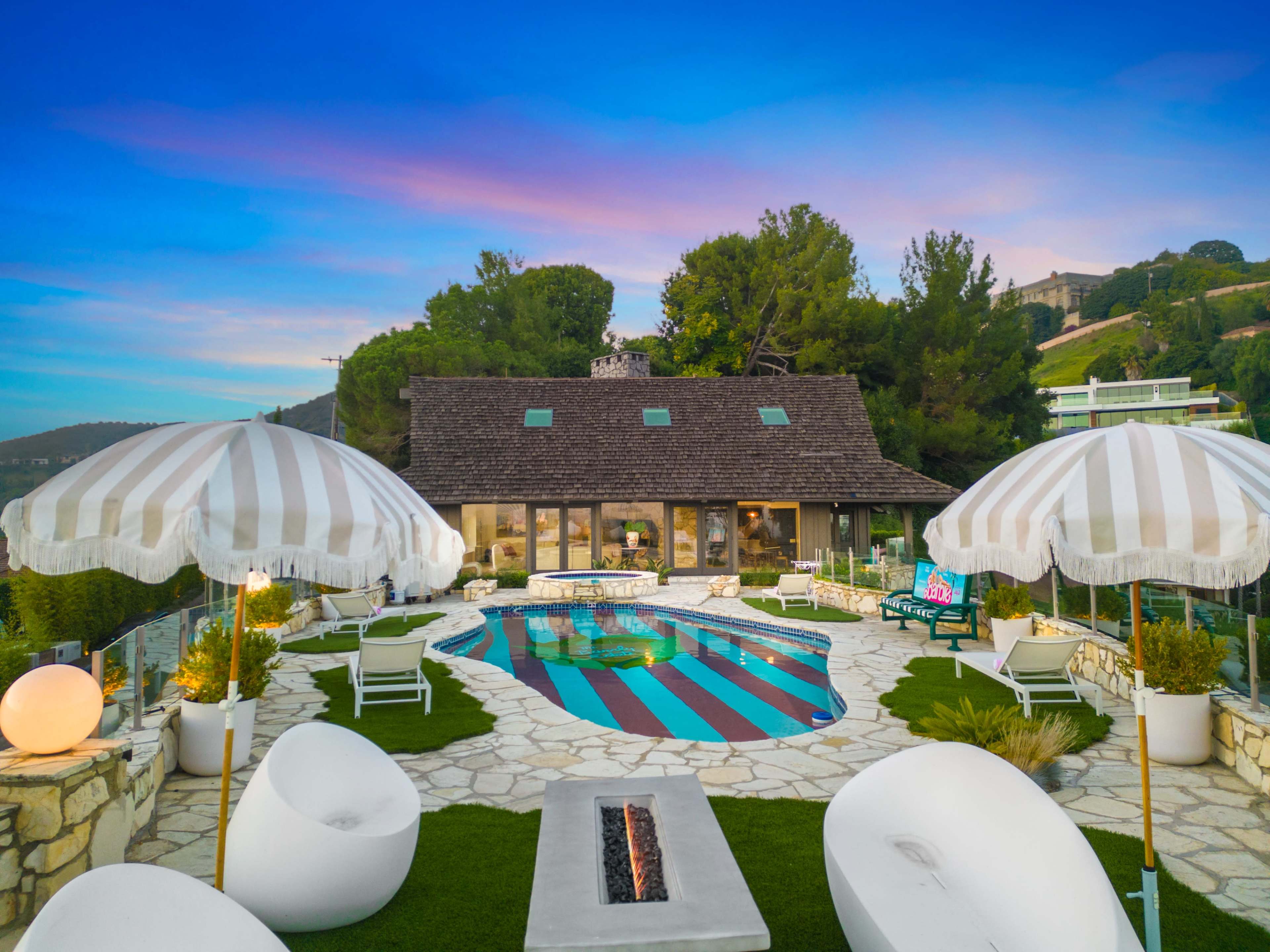 A modern house featuring a pool surrounded by striped umbrellas and lounge seating, set against a backdrop of trees and a sunset sky.