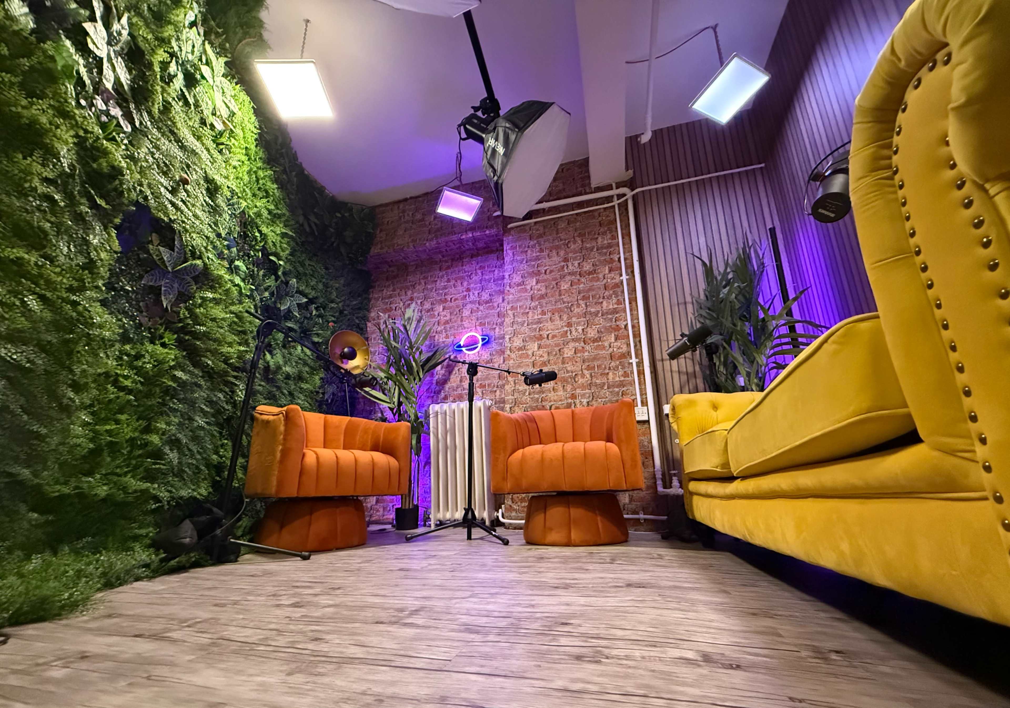 The image shows a modern interior space with orange and yellow sofas, a brick wall, green plants, and studio lighting.