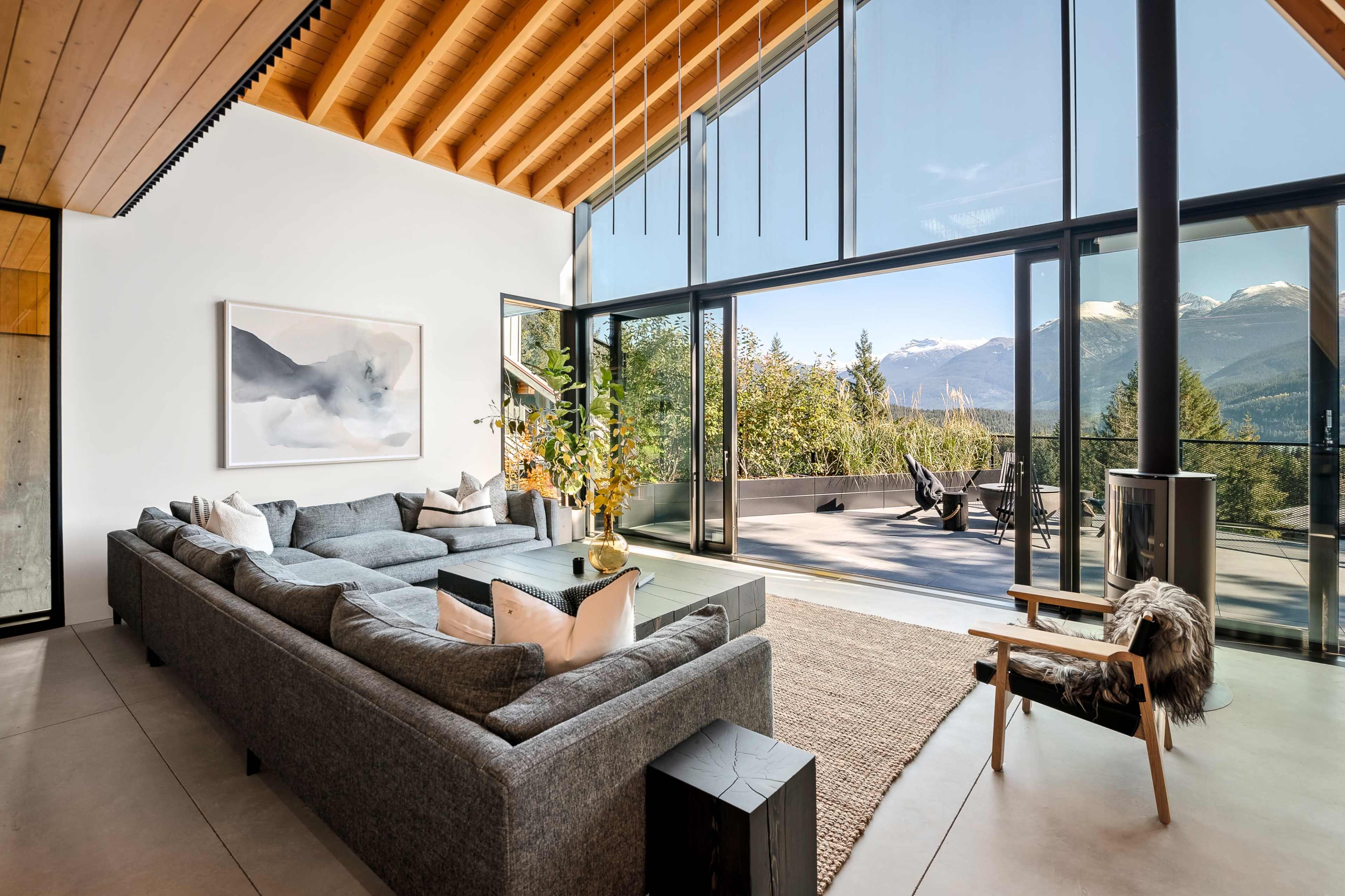 A modern living room features a large gray sectional sofa, panoramic windows showcasing mountain views, and a sliding glass door that opens to a spacious outdoor terrace.