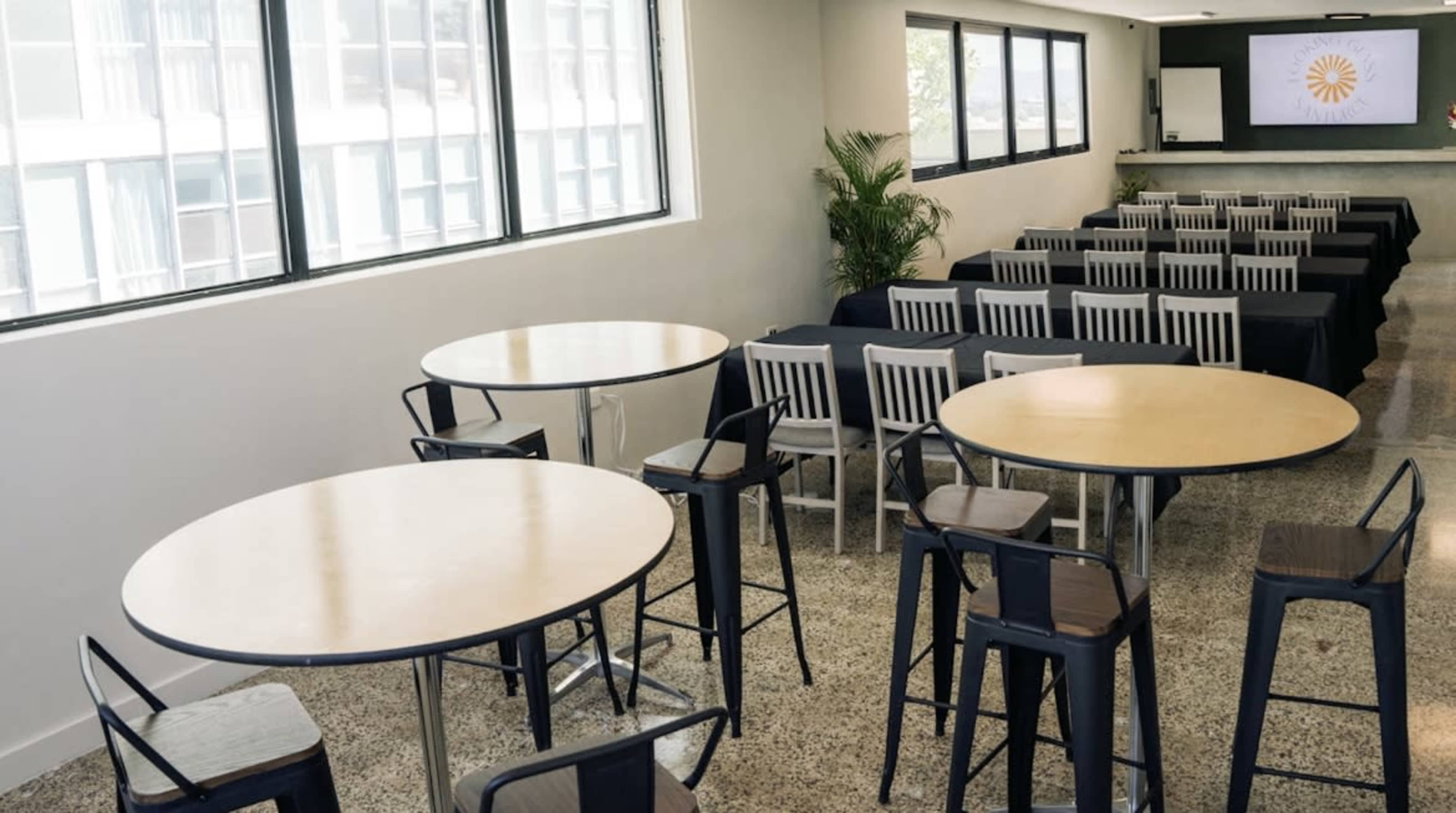 The image shows a modern conference room with round tables in the foreground and rows of chairs arranged behind them, facing a screen at the front.