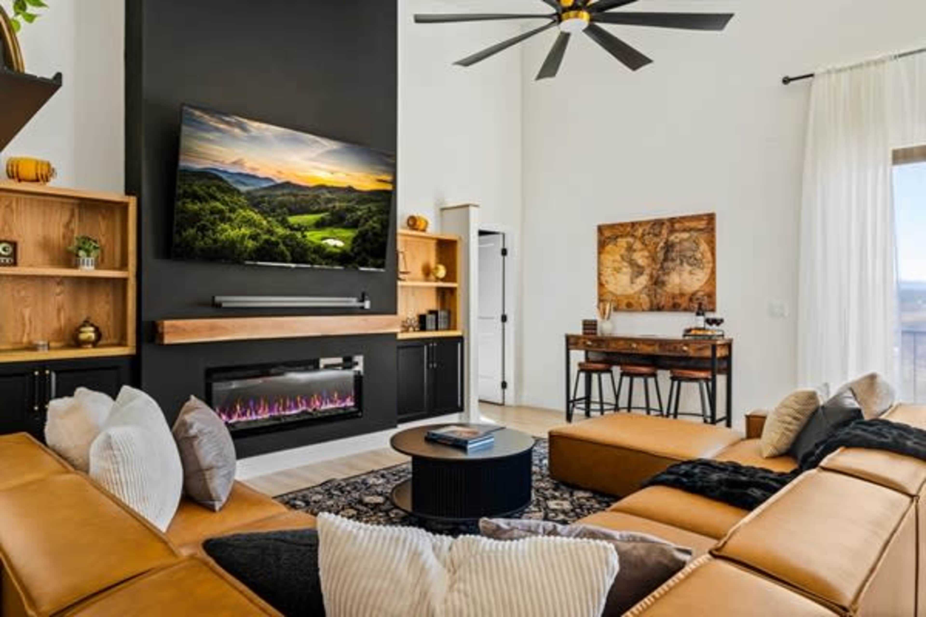 The living room features a wall-mounted television above a modern electric fireplace, flanked by wooden shelves, a sectional sofa, and a dining table with chairs in a well-lit space.