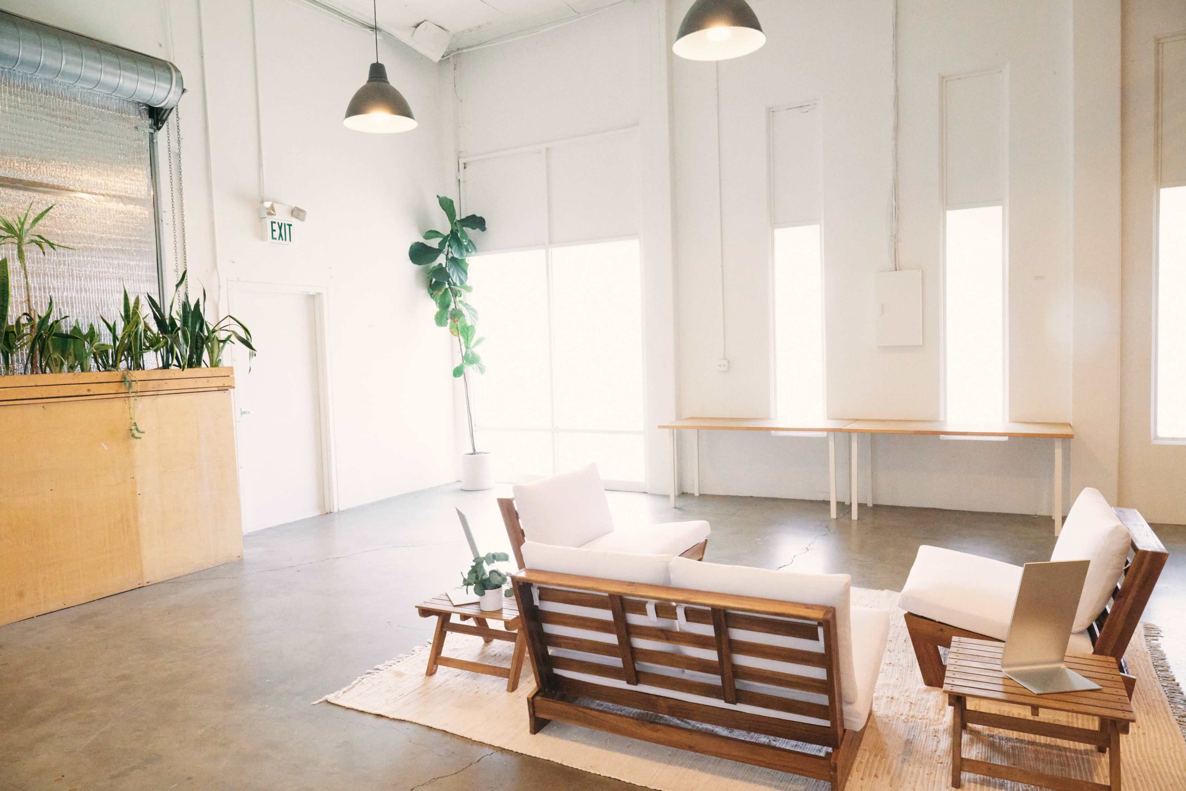 A bright, minimalistic interior features a seating arrangement with white couches and a wooden table, surrounded by large windows and indoor plants.