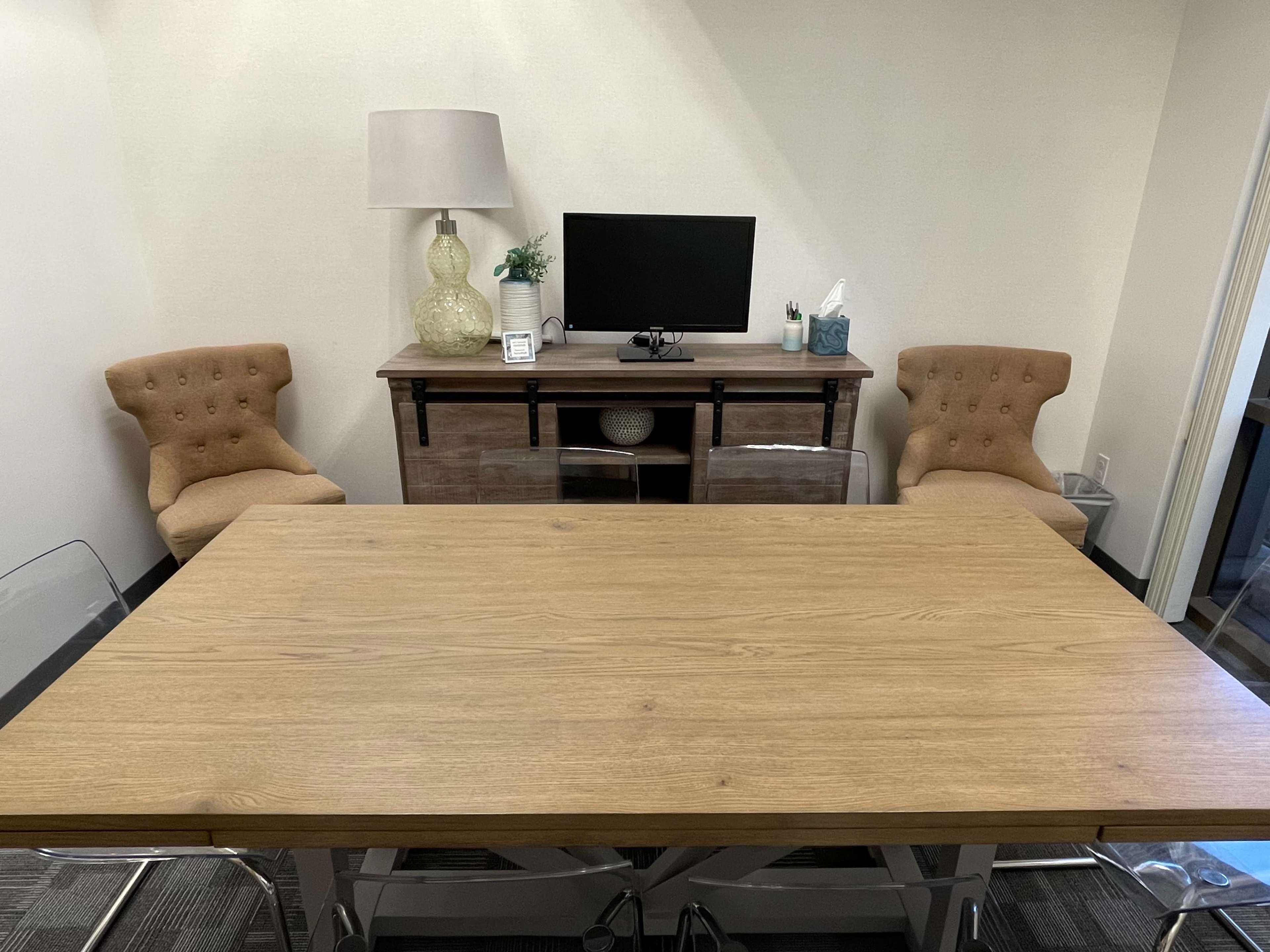 A modern conference room features a wooden table, two upholstered chairs, a lamp, a television on a cabinet, and a tissue box on the surface.