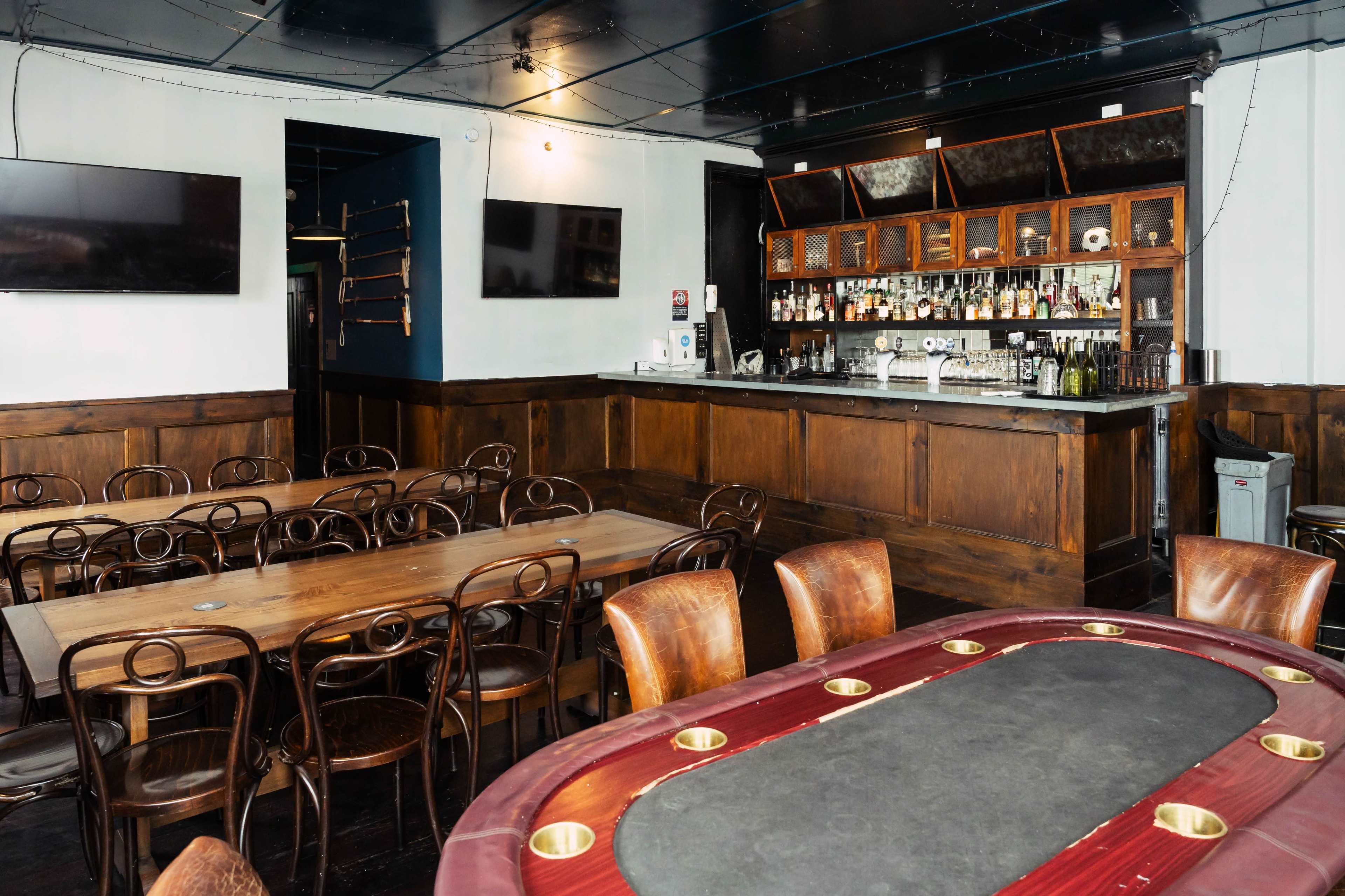The image shows a dimly lit bar interior with a polished wooden bar, shelves filled with bottles, and several tables and chairs arranged around the space.