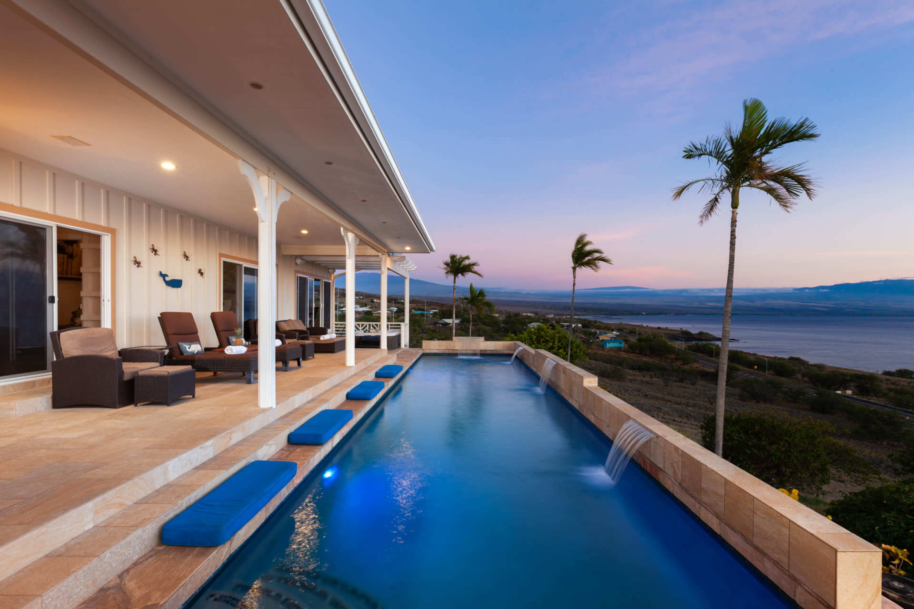 A modern home features a sleek infinity pool with lounge chairs and palm trees, overlooking a scenic view at dusk.