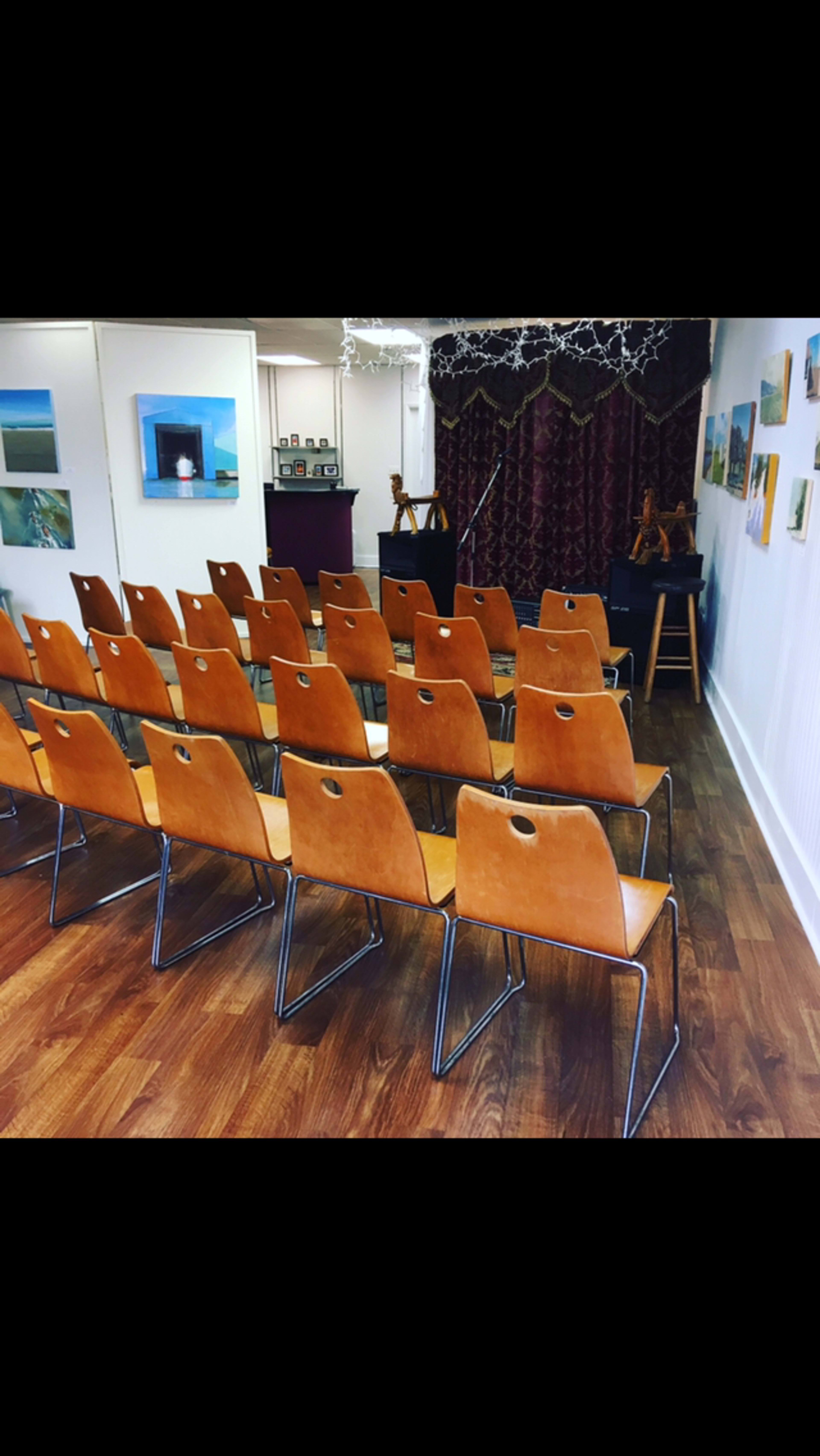A row of stacked brown chairs arranged in a small, decorated performance space with artwork on the walls.