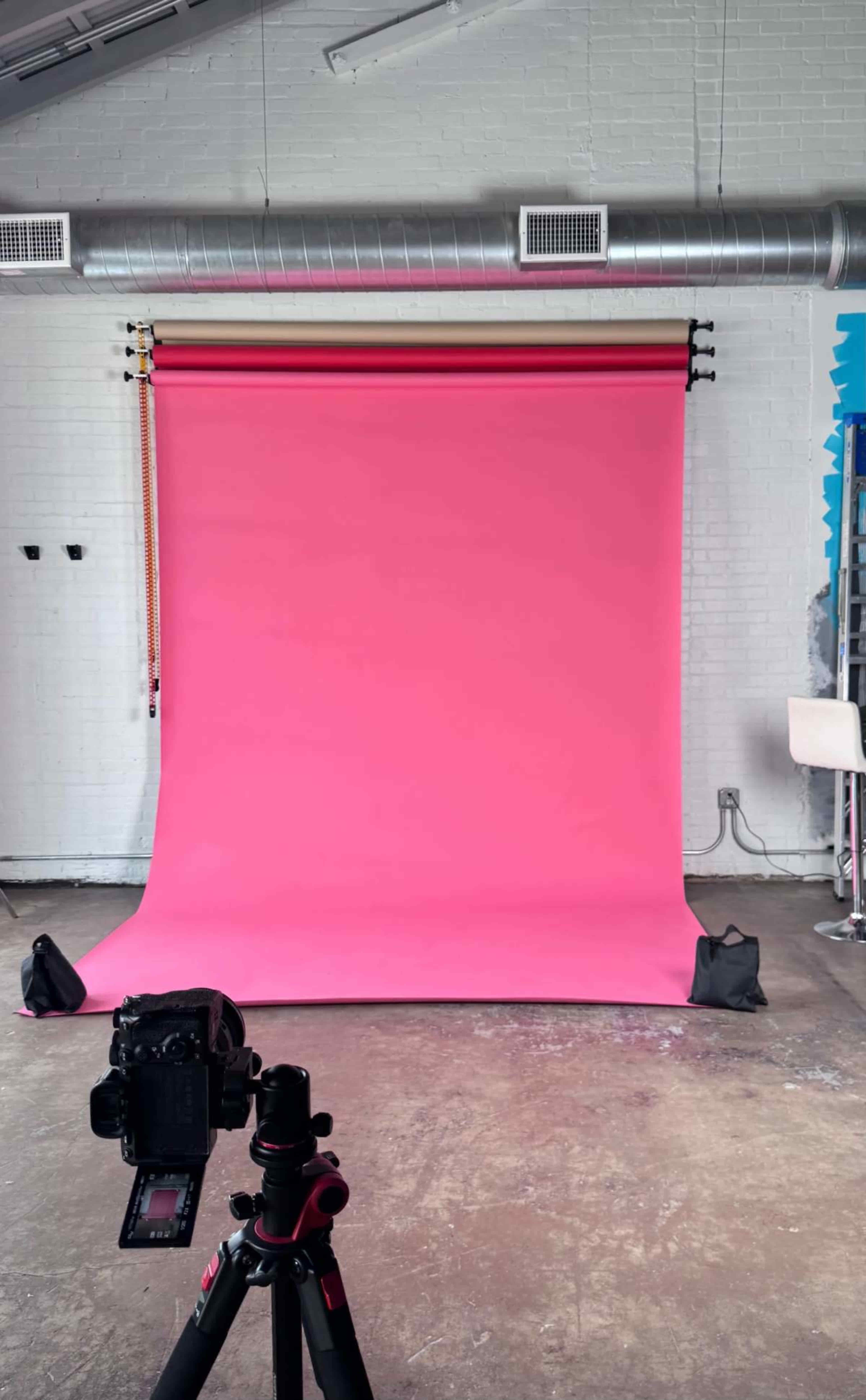 The image shows a photography studio with a bright pink backdrop and a camera on a tripod positioned in front of it.