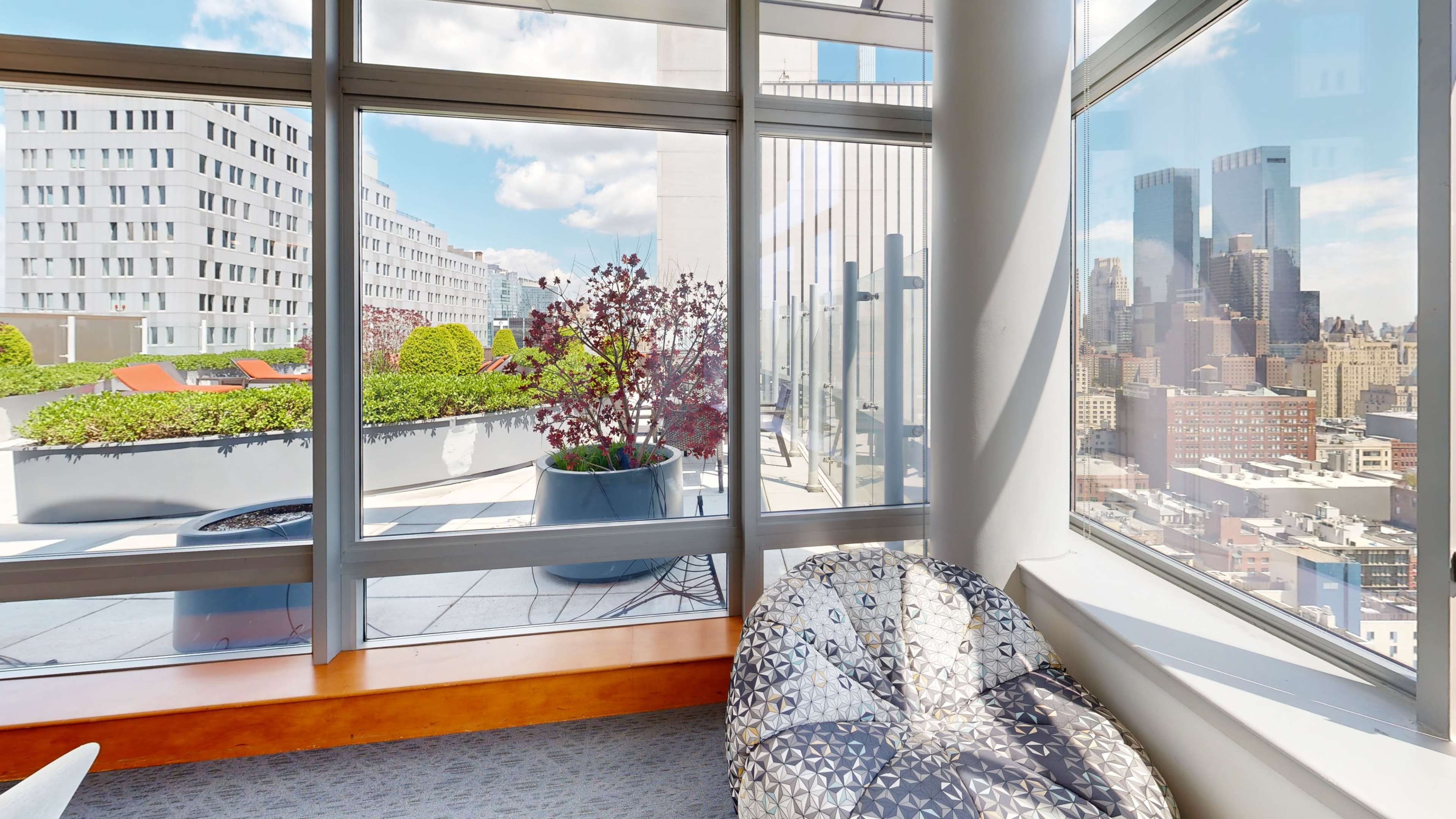 The image shows a modern interior with large windows that provide a view of a rooftop garden and the skyline of a city in the background.