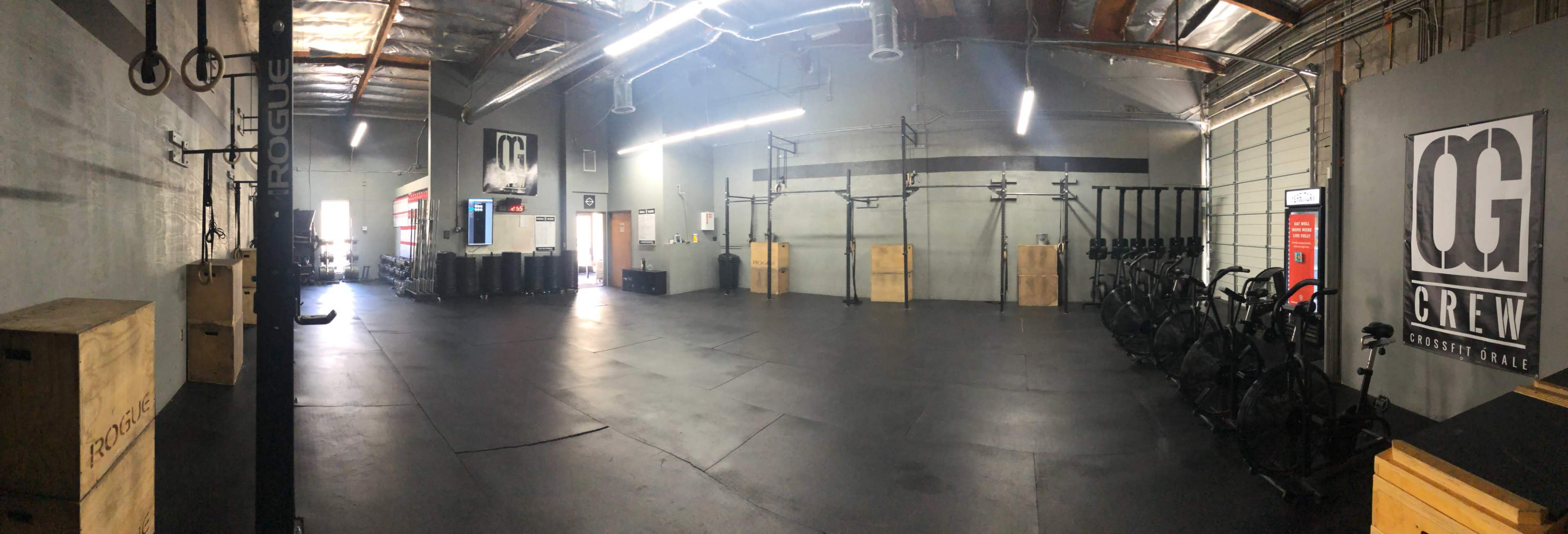 A spacious gym interior with various workout equipment, including rowing machines and wooden boxes, set on a black rubber floor under bright ceiling lights.