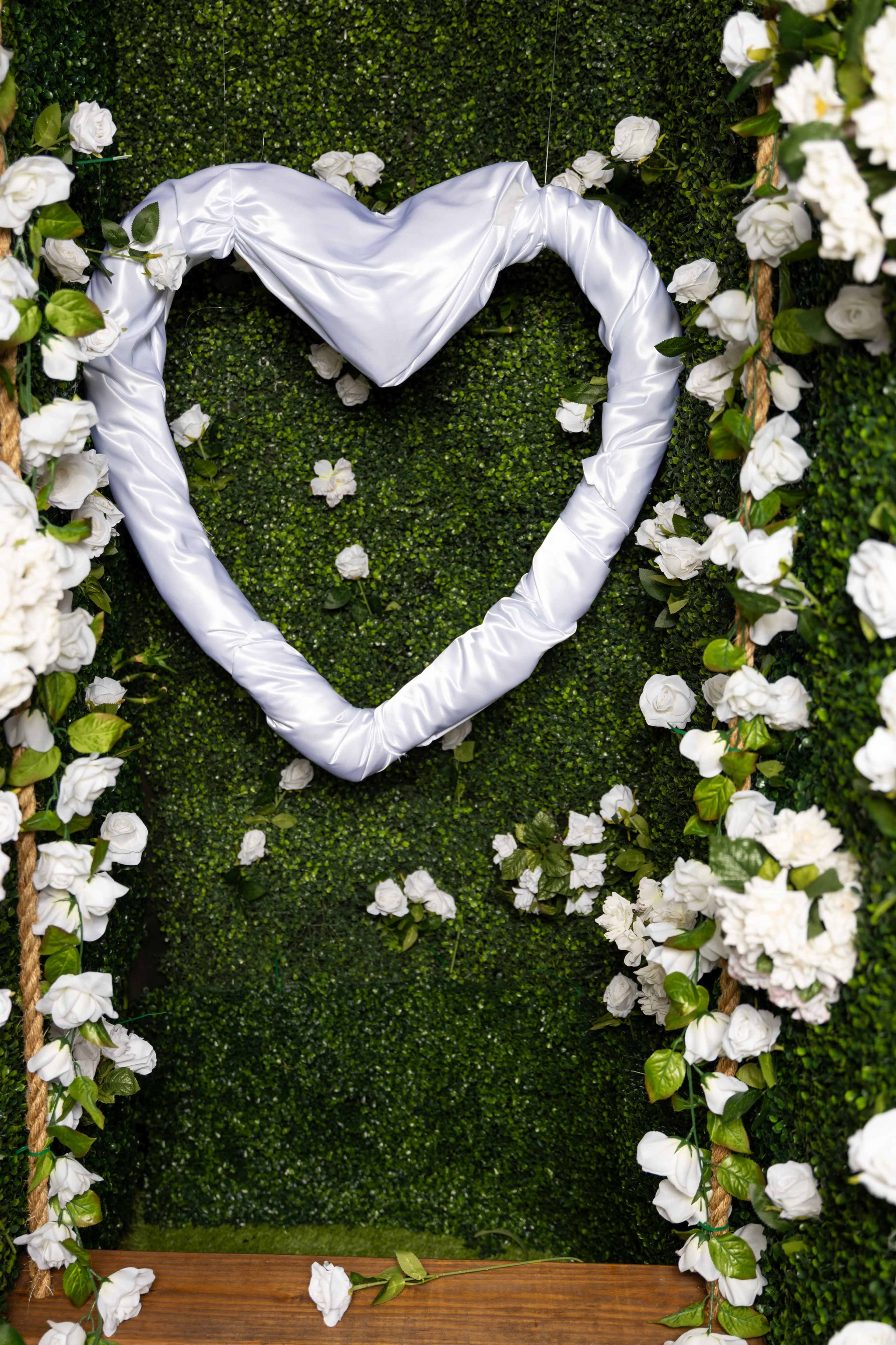 The image features a heart-shaped decoration made of white fabric surrounded by artificial flowers on a green textured background.