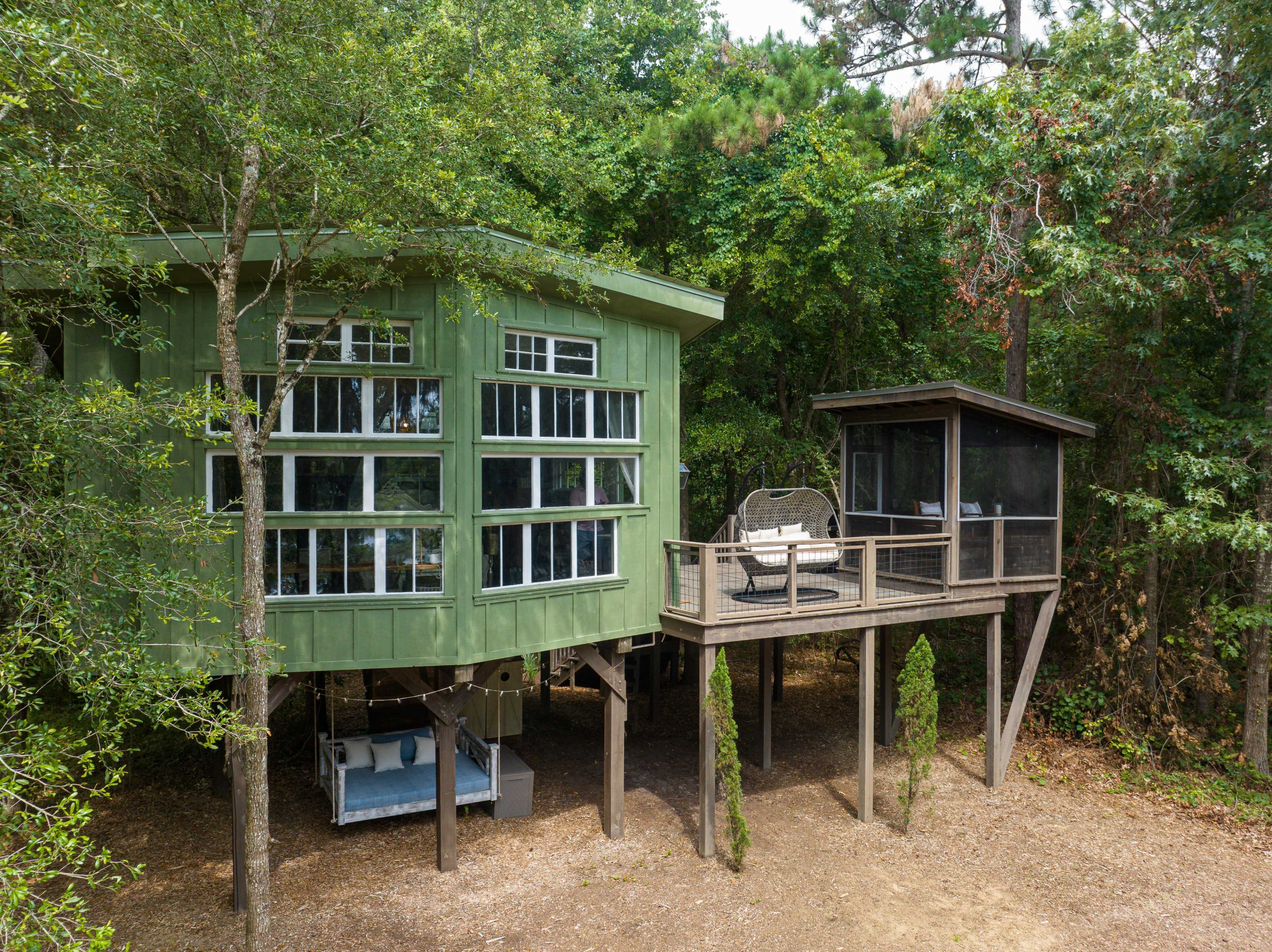 A green, two-story treehouse with multiple windows sits elevated on stilts in a wooded area, featuring a deck with seating.