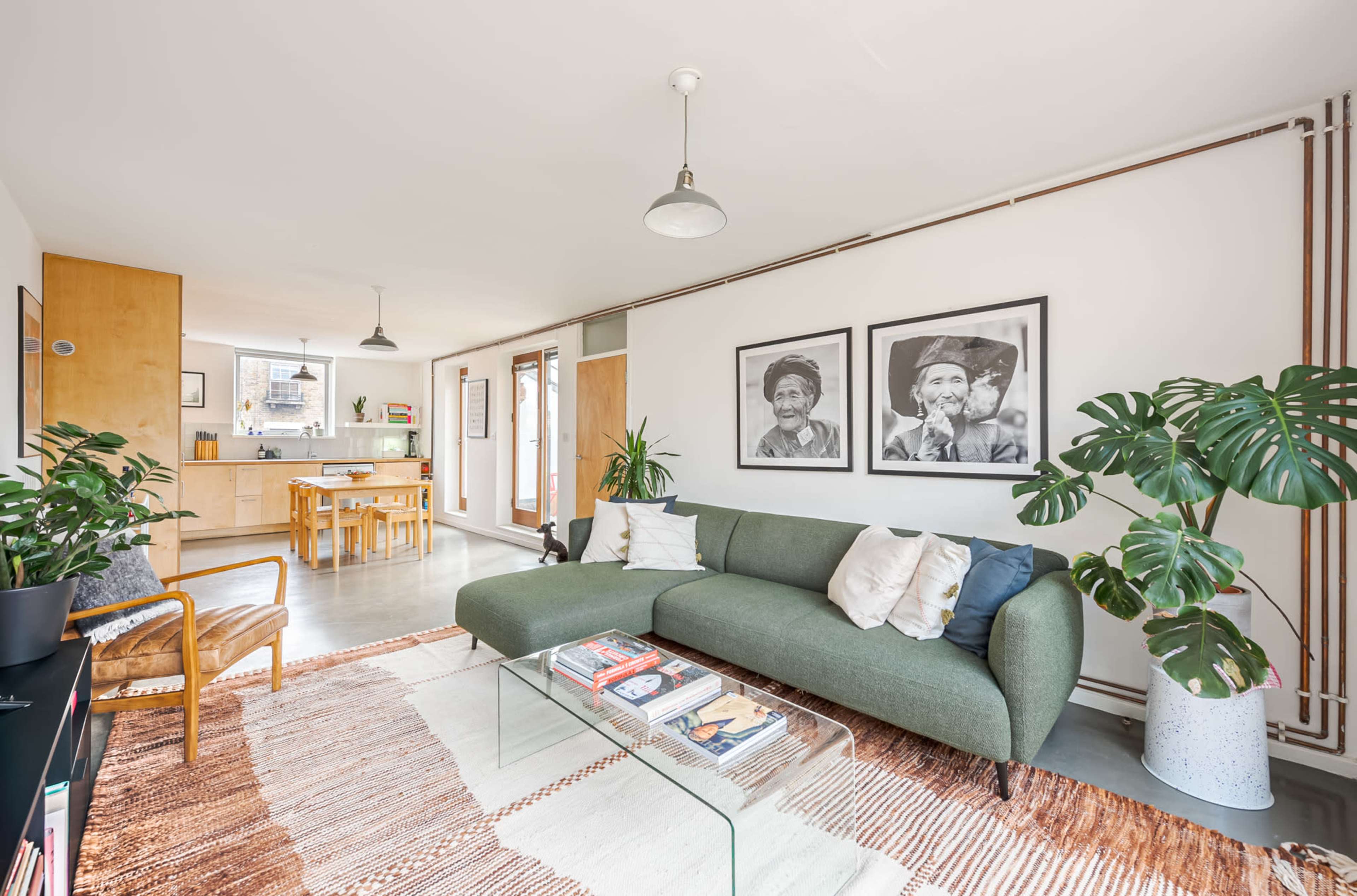 A modern living room with a green sofa, a glass coffee table, and framed black-and-white photographs on the wall, adjacent to an open kitchen area.