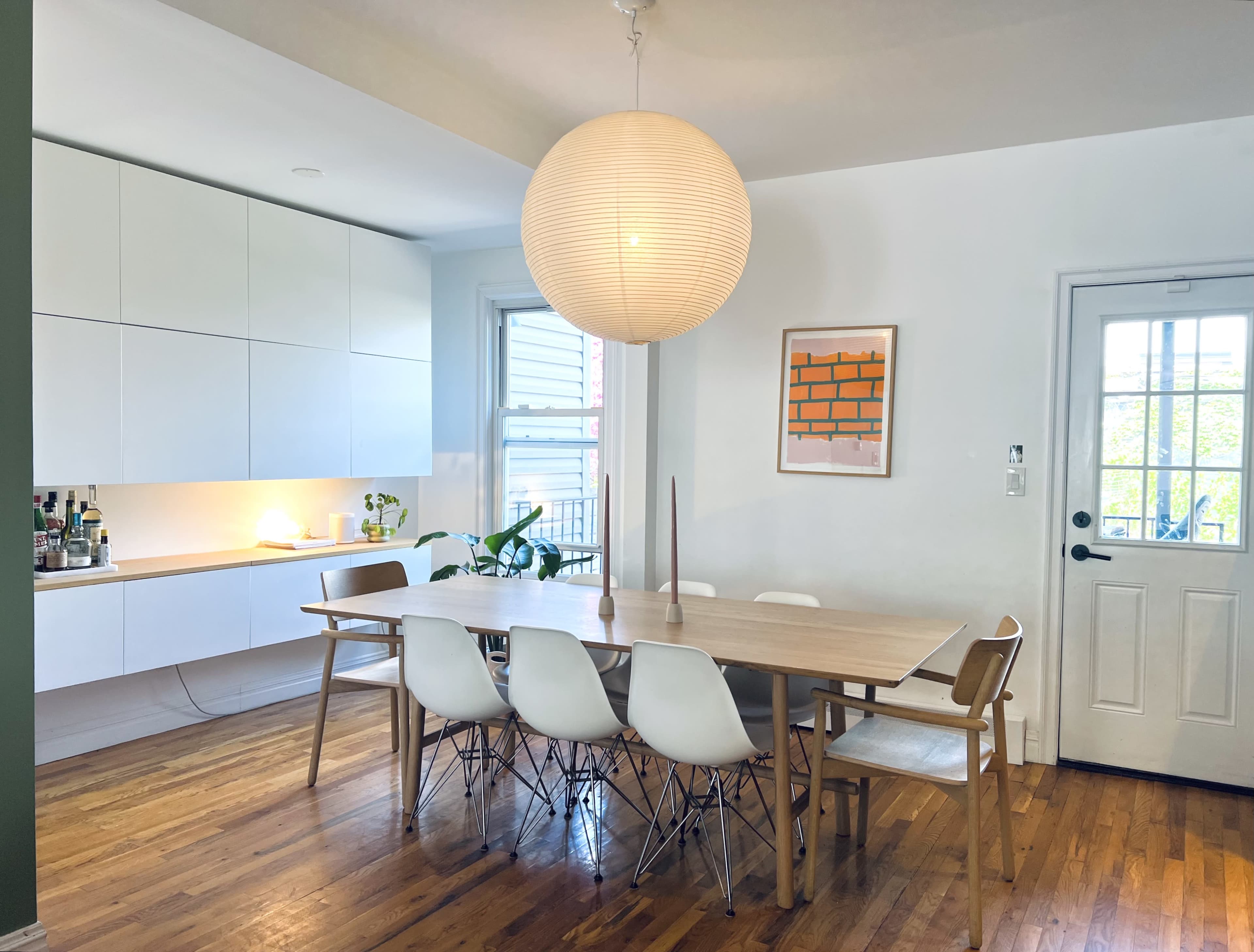 A dining area features a wooden table surrounded by white chairs, with a large pendant light overhead and a view of a door leading outside.