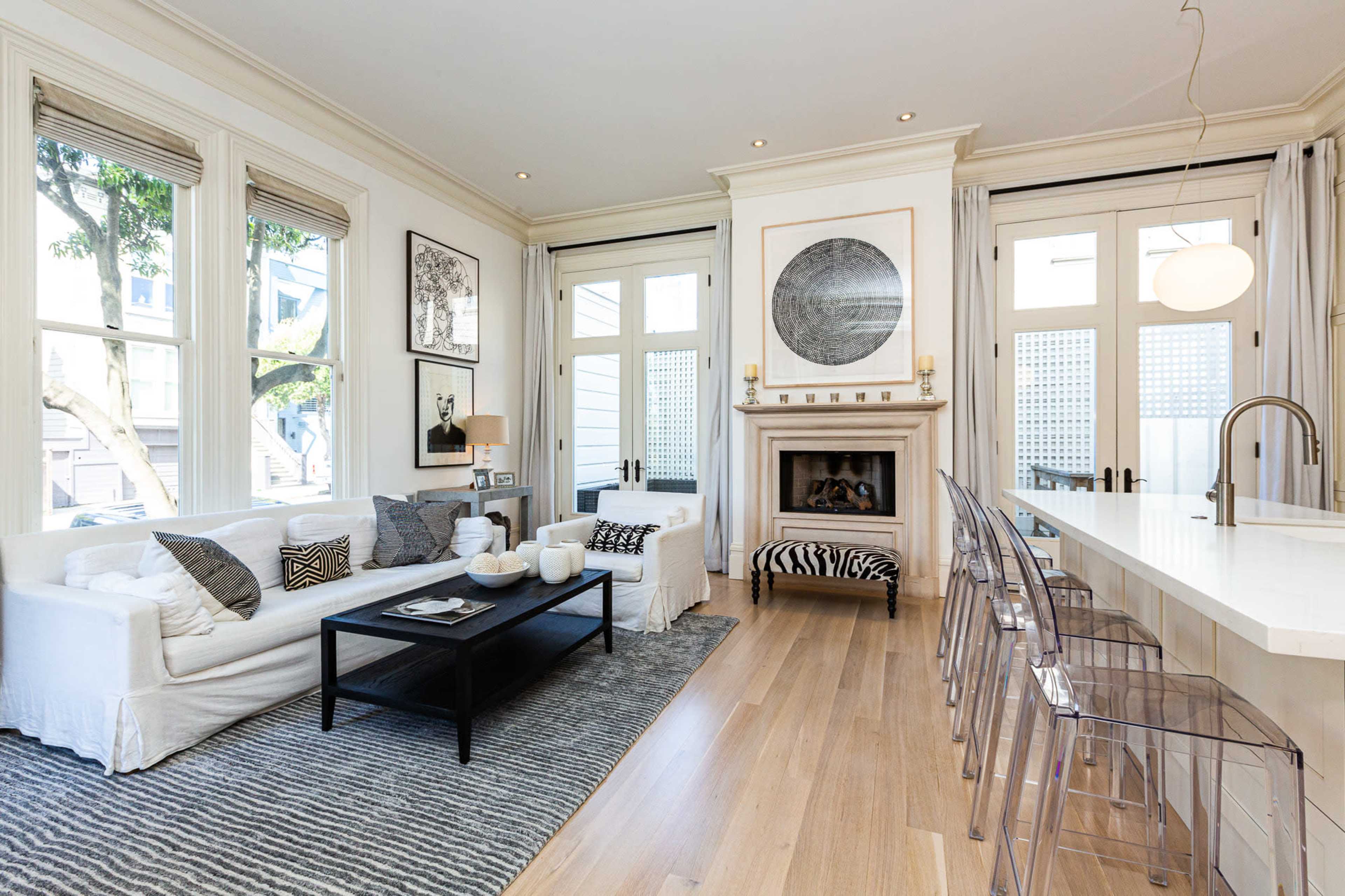 The image shows a modern living room with a white sofa, a black coffee table, and a kitchen bar featuring transparent chairs, all illuminated by natural light from large windows.